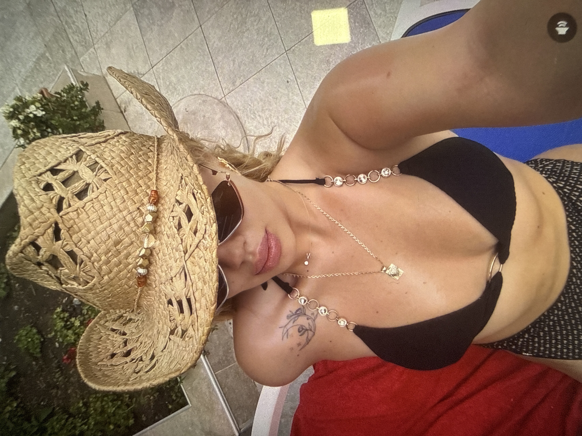 A woman wearing a straw cowboy hat, sunglasses, and a black bikini poses for a selfie outdoors, likely at a resort in Rakovski, Bulgaria. She is lying down on a lounge chair, with a red towel to her left and a tiled floor visible in the background. She has a tattoo on her left shoulder and is wearing necklaces and earrings. The scene suggests a relaxed, sunny atmosphere.