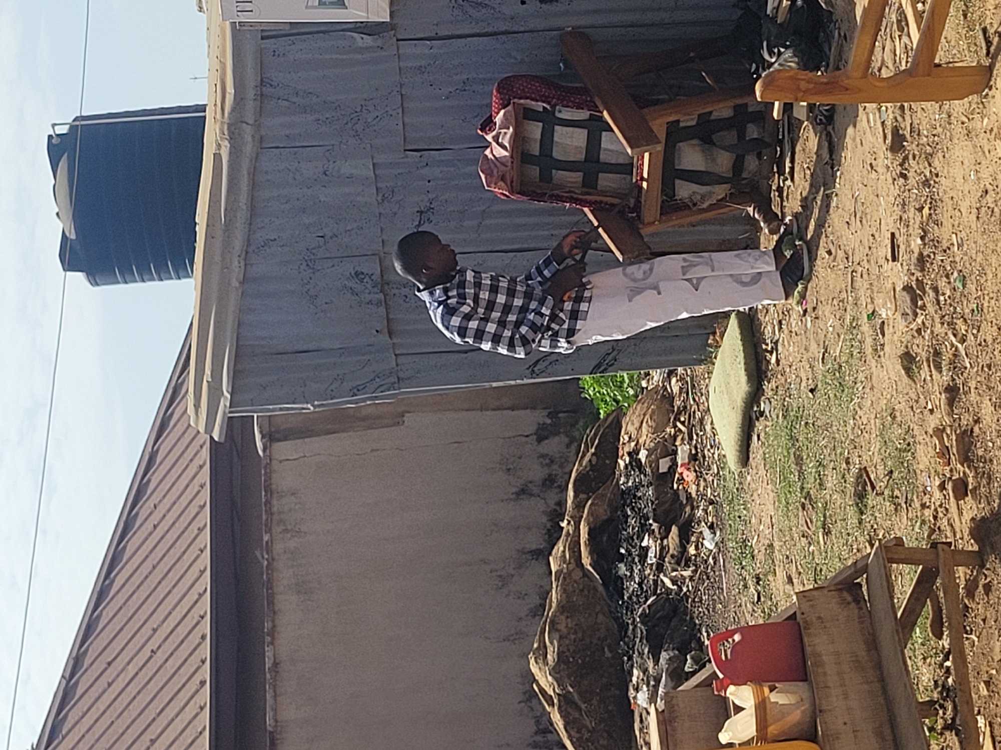 The scene depicts an individual sitting on a chair outside a structure with corrugated metal siding in what appears to be Jos, Nigeria. A black water tank is visible atop a building. The person is wearing a checkered shirt and white pants. There is debris and vegetation on the ground. Adjacent to the structure with the person sitting is a building with a brown roof.