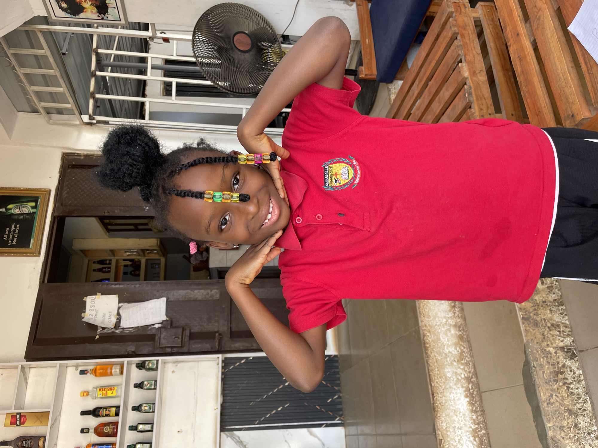 A cheerful young girl is featured prominently, posing playfully with her hands framing her face and a wide smile. She has dark skin and her braided hair, adorned with colorful beads (yellow, green, orange, purple) along the strands framing her face, is pulled into a high bun. She wears a red short-sleeved polo shirt with an embroidered crest on the left chest and black athletic pants with a white stripe down the side.
The setting appears to be an indoor or semi-outdoor area in Barakin Akawo, Nigeria, likely during the daytime. In the background, to the upper right, a dark-colored ceiling fan is visible. Below it are stacked wooden pallets or furniture. To the left, shelves stocked with various bottles, resembling alcoholic beverages, are seen. Brands like "LONG RIDER" and "CHELSEA" are discernible on some bottles. A framed picture showing a person with dark hair hangs on the wall above these shelves, and another framed item carries the text "All men are born equal. Not all beers." A dark brown door is partially visible in the middle background, with a white piece of paper taped to it displaying handwritten numbers and words. The floor is tiled.
Visible text includes:
- On the girl's shirt logo: "RHEMAVILLE", "CHRISTASAUGENT", and "Education for Life".
- On a framed item: "All men are born equal. Not all beers."
- On bottles: "LONG RIDER" and "CHELSEA".
- On the paper on the door: "32 88032", "POT", and "GRACE".