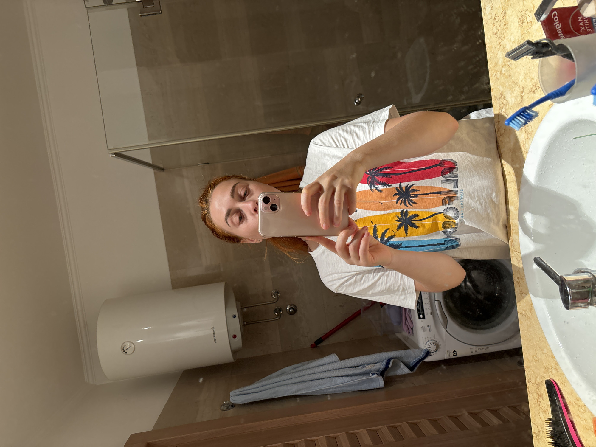 A woman is taking a selfie in a bathroom. The bathroom contains a shower with a glass door, a sink with various toiletries, a water heater mounted on the wall, and a washing machine. The woman has red hair and is wearing a white t-shirt with a design of surfboards and palm trees. The location may be in Budva, Montenegro, based on the style of the bathroom fixtures and appliances, which are commonly found in that area.