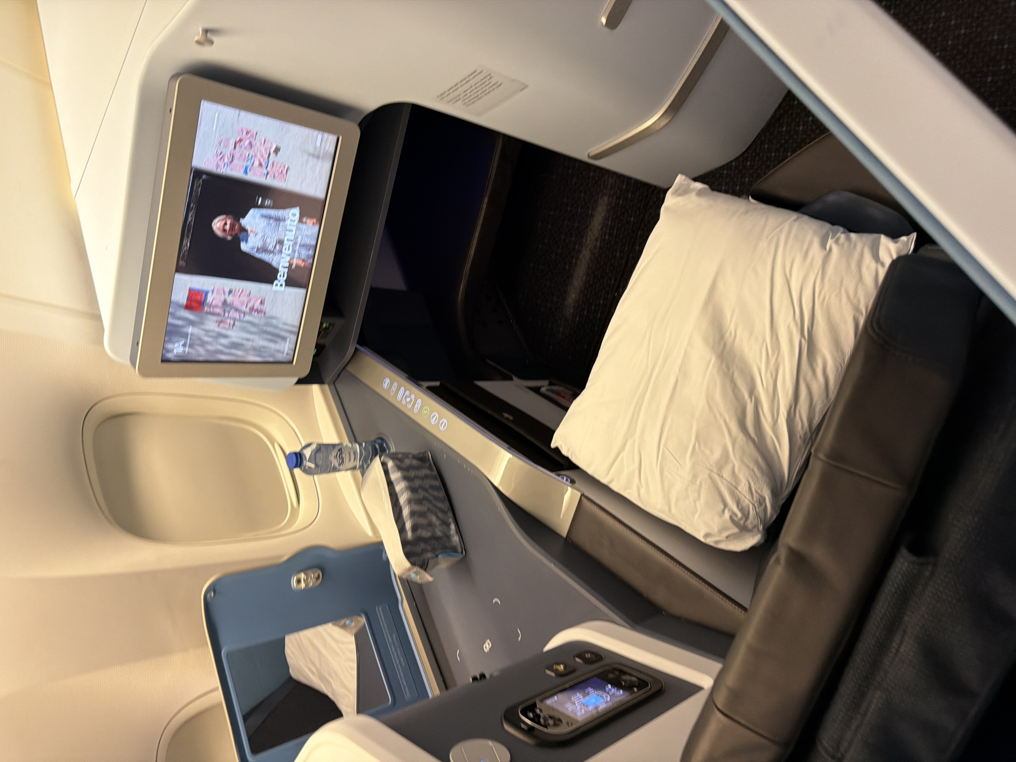 The image depicts the interior of a business class airplane seat. The seat is reclined and features a dark leather armrest. A white pillow is placed on the seat. To the left of the seat, there is a dark grey surface with a built-in screen displaying a person, and what appears to be a control panel. A bottle of water and a toiletries bag are also present on this surface. The background shows a window and the wall of the airplane cabin. The scene is well-lit, suggesting it is inside the cabin during daytime or with artificial lighting. This appears to be a static, posed shot rather than an action shot. There are no people visible in the image. The location within Schiphol, Netherlands is not discernible from the image itself.