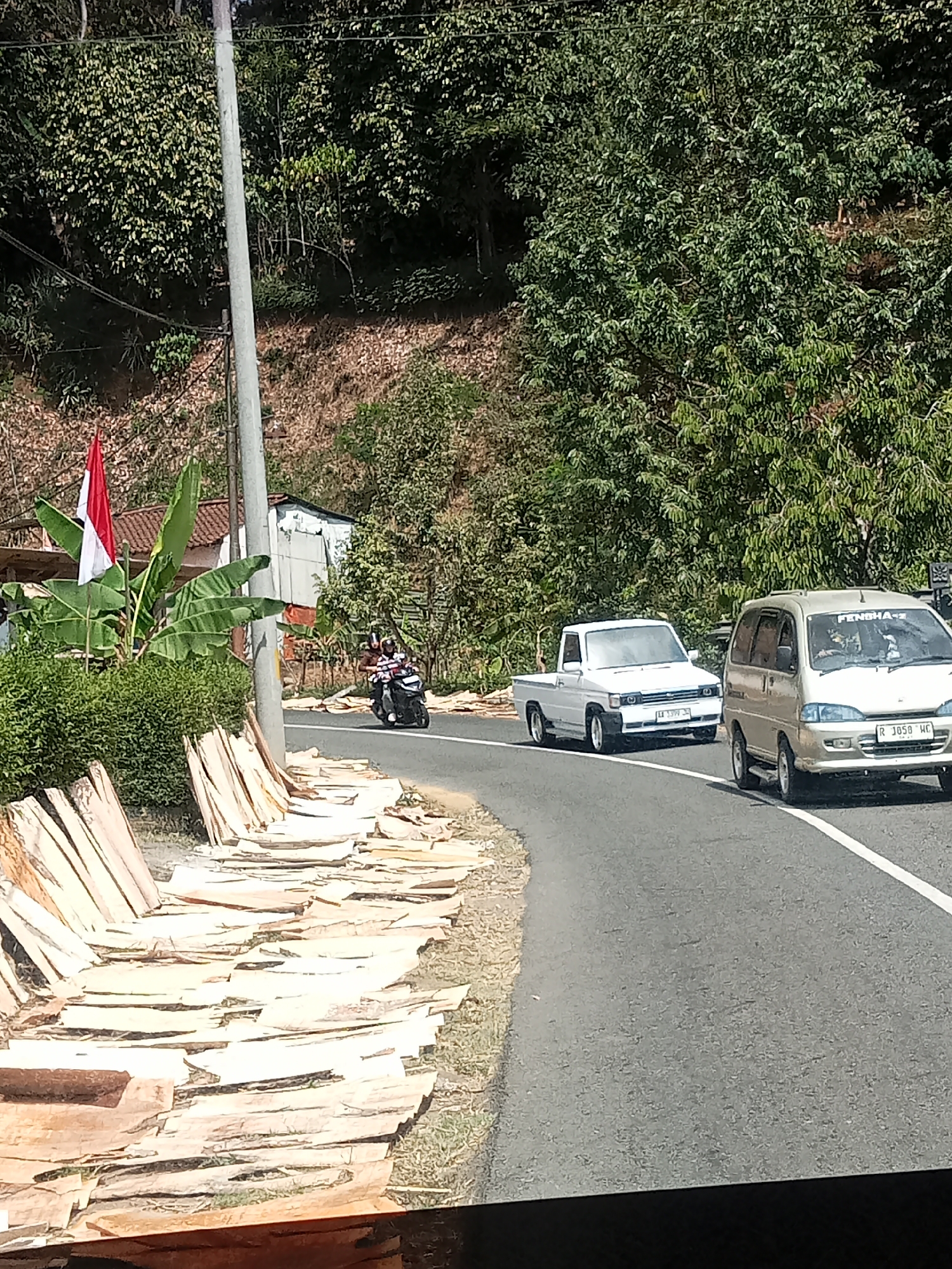 In Gambarsari, Indonesia, a paved road curves through a vegetated hillside. On the left side of the road, large stacks of light-colored, unprocessed wood are laid out on the ground. Further up the embankment, a small structure with a white facade and a tiled roof is visible, with a red and white Indonesian flag planted nearby, alongside lush green banana leaves.
A motorcycle with two occupants rides along the road, heading away from the viewer. In the background, a white pickup truck and a beige van are traveling in the same direction as the motorcycle. The pickup truck has a license plate that reads "AA 1399 JP," and the van's license plate is "R 3050 HE." The van also displays the word "FENSHA" on its windshield. The environment is characterized by dense green foliage covering the hillsides and a clear sky overhead.