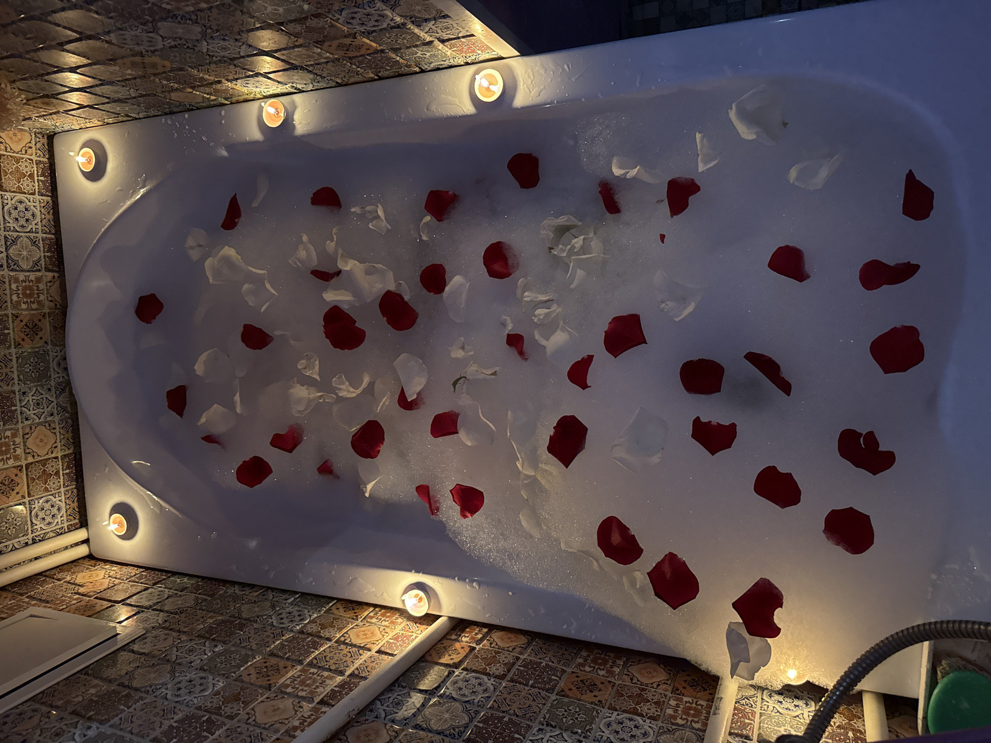 A top-down, slightly angled view shows a bathtub filled with soapy water and scattered red and white rose petals. Six small candles are placed along the rim of the tub. The surrounding floor and wall are covered in patterned tiles. A shower head is visible in the lower right corner of the frame. The location is implied to be in a bathroom in Chaikovsky, Russia, but there is no directly visible evidence to confirm this. There are no people visible in the image.