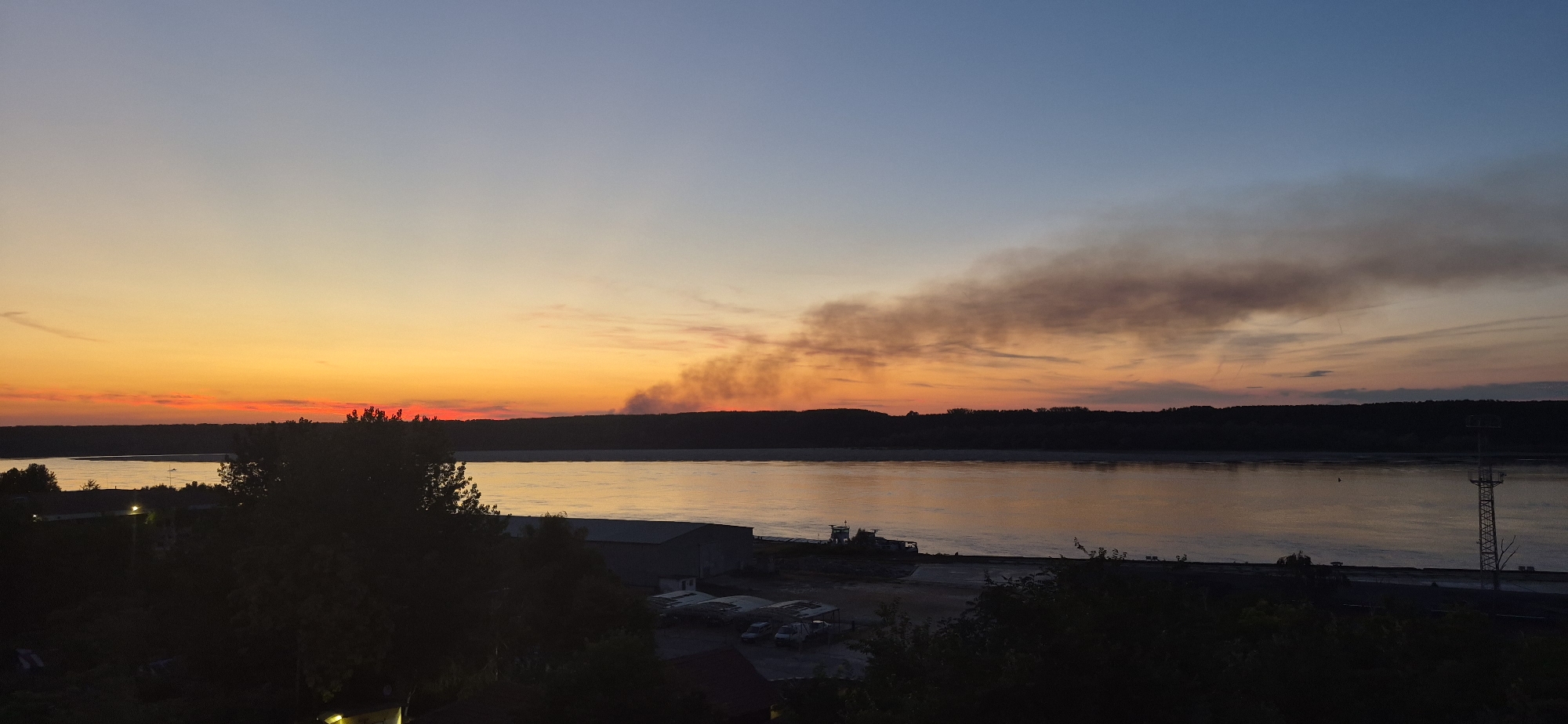 A wide shot of a river at sunset, with a dark silhouette of trees and buildings on the banks. A plume of smoke rises from the far bank, partially obscuring the sunset. The sky transitions from a bright orange near the horizon to a pale blue at the top. The river reflects the warm colors of the sky. On the near bank, several industrial-looking buildings and parked vehicles are visible in silhouette. A tall, metal tower stands on the far right bank. The scene is likely taken from an elevated position overlooking the river.