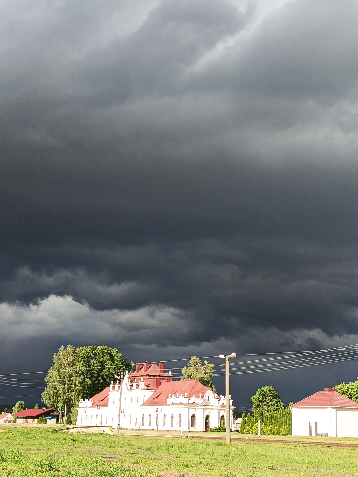 The image depicts a cloudy sky over a rural landscape in Лосева, Belarus. Dark, ominous storm clouds fill most of the frame. Below the clouds, a low-lying green field stretches across the foreground. In the middle ground, several white buildings with red roofs are visible. A line of utility poles and wires cuts across the scene. Trees are interspersed among the buildings. There are no people visible.