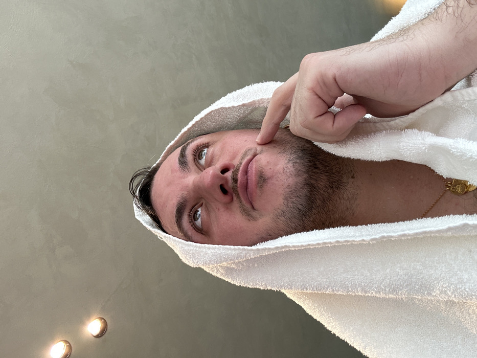 A male subject is depicted in an indoor setting, viewed from a low angle, with his head and upper torso visible. He has short, dark, possibly wet hair, and facial stubble on his cheeks and chin, with a mustache. He is wrapped in a white terry cloth towel, draped over his head and shoulders like a hood. His gaze is directed upwards. His right index finger is touching his right cheek/jawline. A gold chain with a gold-colored, possibly religious, pendant is visible around his neck. The ceiling above him is a light grey with a textured, mottled finish. Two illuminated recessed circular light fixtures are visible on the ceiling. The specific location within the city of London, United Kingdom, cannot be determined from the visual information presented in the image.
