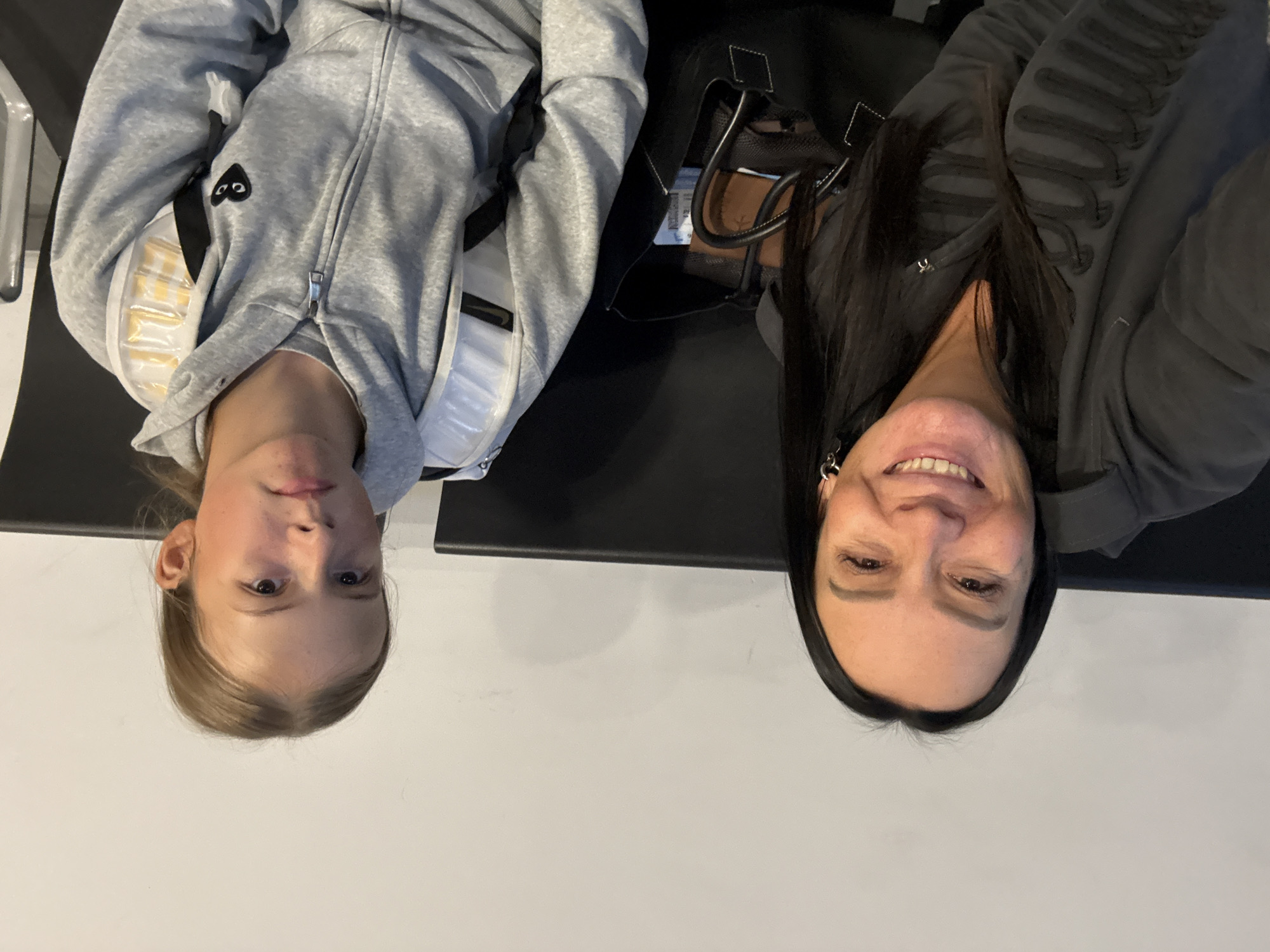 The image shows two females, an adult and a child, seated on a black bench against a plain white wall. The adult, on the left, has long dark hair and is wearing a dark gray jacket with decorative elements. She is smiling at the camera. The child, on the right, has light brown hair and is wearing a gray hooded jacket with a backpack. The child is looking forward, appearing contemplative. A black bag is visible between the two individuals. The overall environment suggests an indoor waiting area, possibly at an airport or train station. The location is identified as Bolshoye Savino, Russia, which is known for its airport.