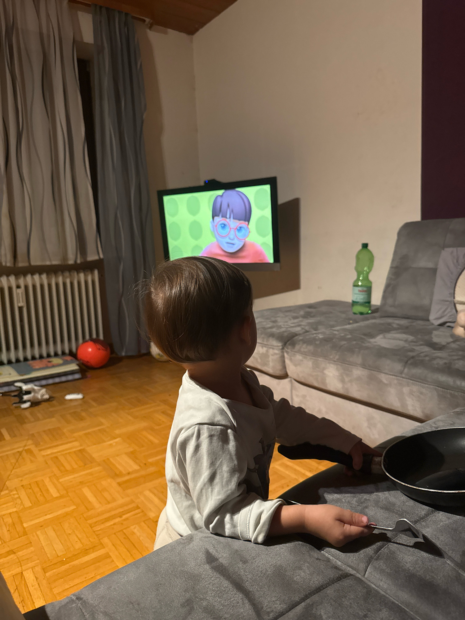 The image depicts a child, seen from behind, watching a cartoon on a television screen. The child is wearing a light-colored shirt and is seated on a gray sofa. In front of the child, a black frying pan and a fork are resting on the sofa. The television displays a cartoon character with large glasses against a green, polka-dotted background. To the left of the television, a window is visible with gray curtains and a white radiator. On the wooden floor, a red ball and some books are scattered. A green plastic bottle is placed on the sofa to the right. The overall scene suggests a domestic setting, likely a living room.