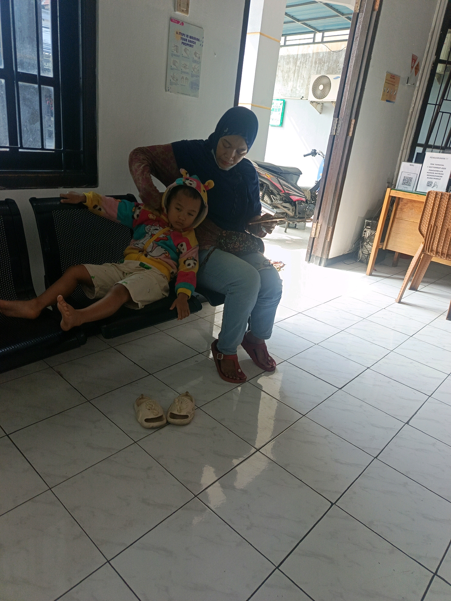 In a waiting area, likely at a clinic or public facility in Sedati Agung, Indonesia, a woman wearing a dark hijab and a patterned top sits on a black bench. She is holding a tablet or phone and looking down at it. Beside her, a young child, dressed in a colorful hooded jacket with animal ears and light-colored shorts, is slumped on the bench, appearing sleepy or bored. The child's feet are bare, and a pair of light-colored sandals with cartoon faces are placed on the tiled floor in front of the bench. The environment features a tiled floor, a large window with dark frames on the left, and a doorway or open passage on the right leading to an outdoor area with a motorcycle and an air conditioning unit visible. Posters are affixed to the wall, one depicting handwashing steps and another with text that appears to be a notice or informational sign. The lighting suggests it is daytime, with sunlight filtering into the space.