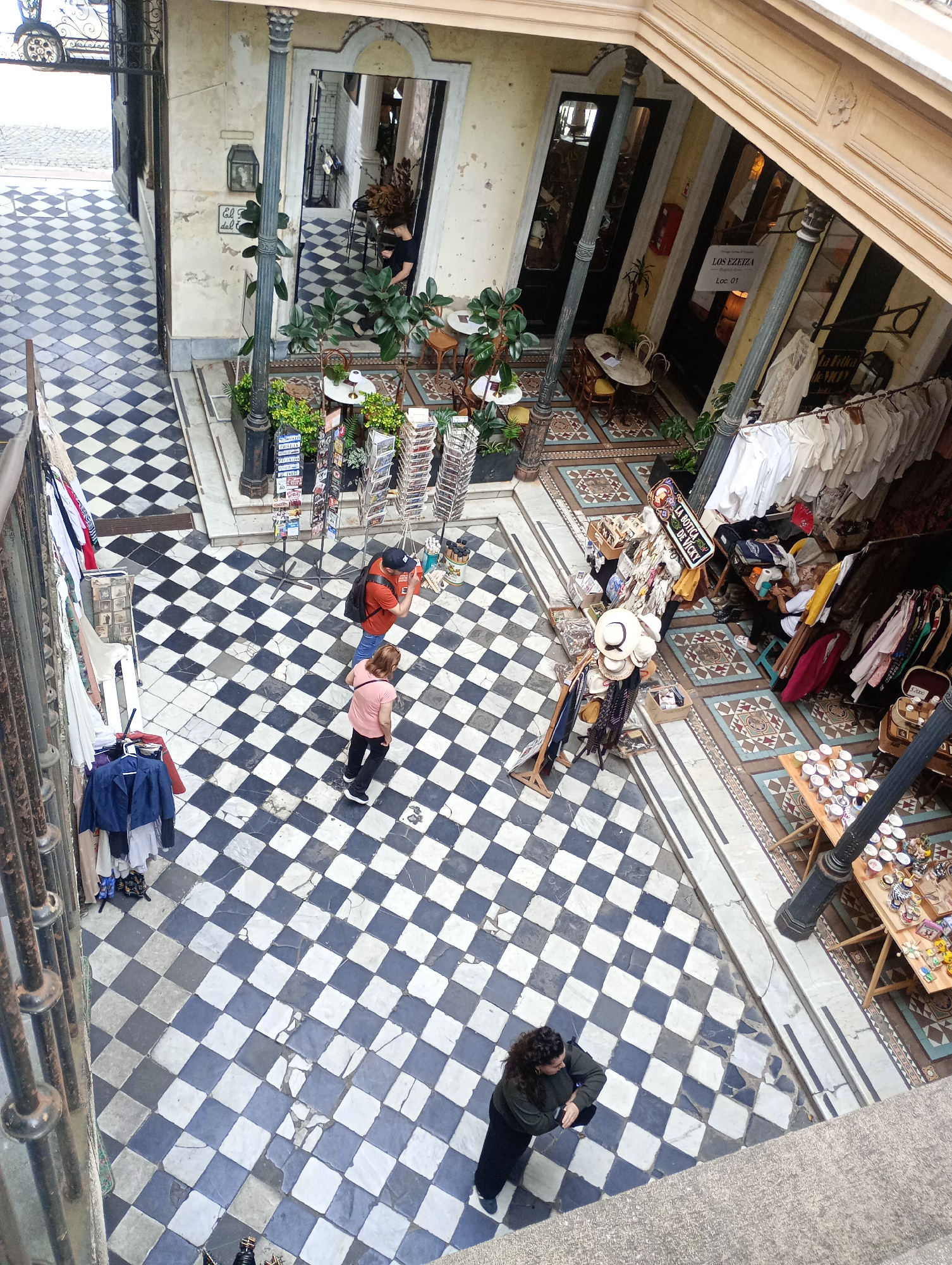 This is an eye-level, outdoor shot of the interior of a retail complex in Buenos Aires, Argentina. The floor is tiled with black and white squares in a checkerboard pattern. Several people are present: two in the middle of the frame, one in an orange shirt and one in a pink shirt. A woman stands near the bottom of the frame. People mill about within individual shops. The shops contain various items for sale, including clothing racks with garments, postcards, hats, and other items on display. Signage for stores such as "LOS EZEIZA" and "LA BOTICA DE VICKY" are visible. Decorative plants and architectural details add to the ambiance of the complex.