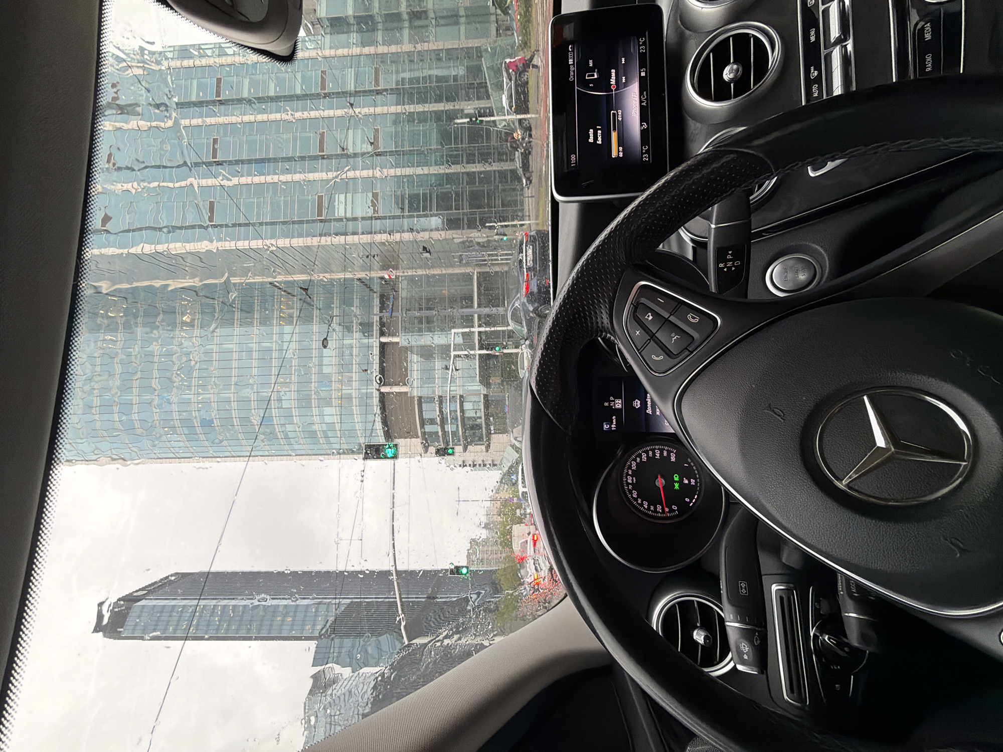 The image captures the view from inside a car, likely a Mercedes-Benz based on the steering wheel and dashboard design. The car is presumably stopped in traffic as indicated by the rain-streaked windshield showing buildings and streetlights in the background. The architecture of the visible buildings is modern, featuring glass and steel structures. The weather appears to be rainy or wet. The dashboard shows the car's systems are active, with a display showing music information, temperature, and car status. Traffic lights are visible through the windshield, indicating an urban intersection.