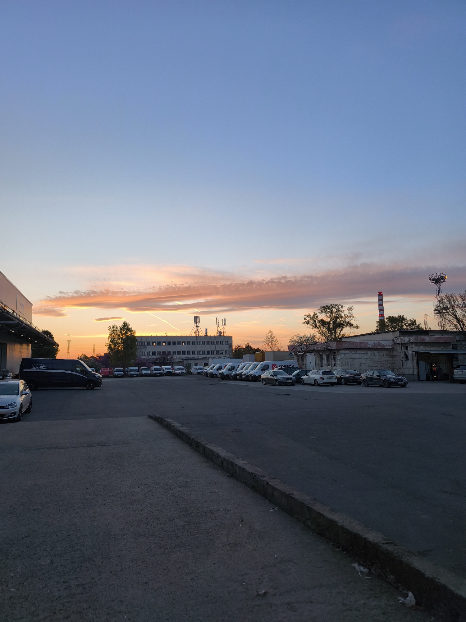 This image depicts an industrial or commercial area in Burgas, Bulgaria, captured during sunset. The sky displays a striking gradient of colors, transitioning from light blue at the zenith to vivid orange and pink hues near the horizon, illuminated by the setting sun. Scattered clouds, some appearing as long streaks, are visible, reflecting the warm light.
The scene is primarily an asphalt parking lot, populated with numerous parked vehicles, including several white and dark-colored vans, alongside various cars. A dark van with a large graphic, possibly resembling a panther, is prominent on the left side of the lot. Further back, a white van clearly displays the "Speedy" logo and name on its side.
In the background, a long, multi-story building with numerous windows, likely an office or factory, stretches across the frame. To its right, a prominent red and white striped industrial chimney rises against the sky, accompanied by a telecommunication tower. Trees are visible scattered throughout the background, between the buildings. On the far left, a portion of a modern-looking building with an extensive roof overhang is visible.
The ground in the foreground consists of rough asphalt, with a distinct concrete curb running along the right side. No people are visible in the image. The overall impression is one of a quiet, functional space at the end of the day, bathed in the dramatic light of the sunset.