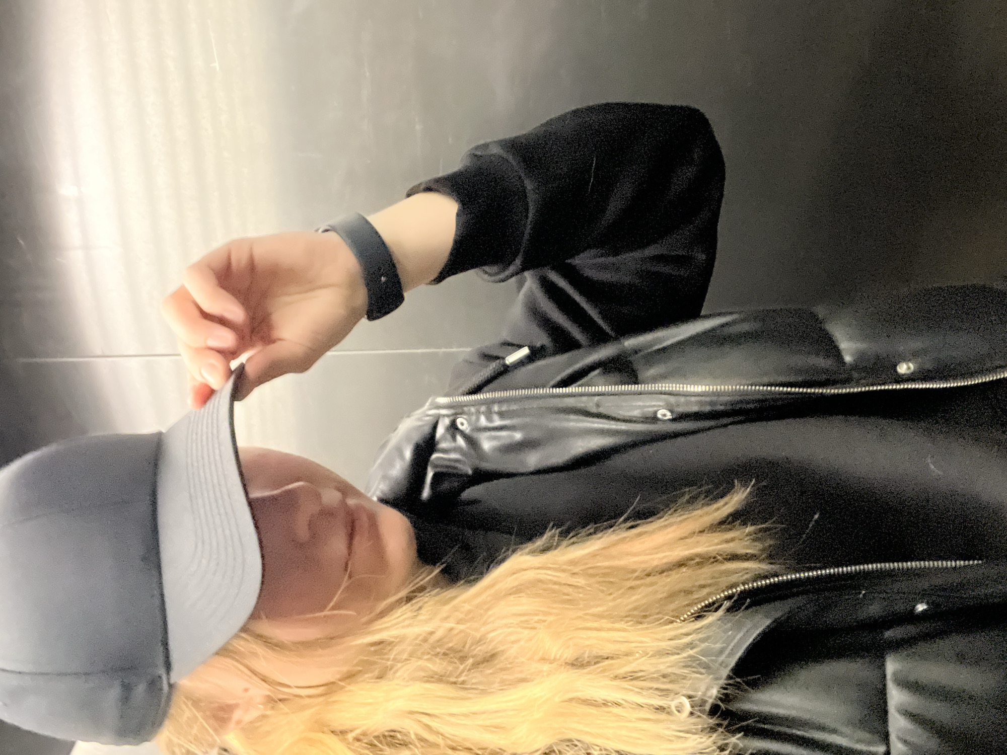 This image is presented in a landscape orientation but depicts a scene that appears to be rotated 90 degrees clockwise from its natural upright position. The central subject is a person, likely female, whose upper body and head are partially visible in a close-up shot.
The individual has long, light blonde or light brown hair that flows towards the bottom of the frame. They are wearing a light grey baseball cap, which their left hand is subtly holding or adjusting near the brim. Their face is largely obscured by the cap and the angle, making their expression undiscernible. The person is dressed in dark, casual attire, including a black hooded sweatshirt and a black puffer vest or jacket. The outerwear features visible silver zippers and snaps, suggesting a faux-leather or shiny material. A dark-colored smartwatch or fitness tracker is worn on their left wrist.
The activity appears to be a casual pose, with the individual perhaps in the process of adjusting their cap.
The background is an indoor setting with two distinct areas. To the left side of the frame (which would be above the person if the image were upright) is a light-colored wall with a subtle vertical striped or textured pattern, possibly metallic or painted. To the right side, behind the person's back, is a darker, plain surface. The lighting is even and diffused, indicative of an indoor environment. There are no specific cues within the image to identify the location as Mytishchi, Russia. No visible text is present.