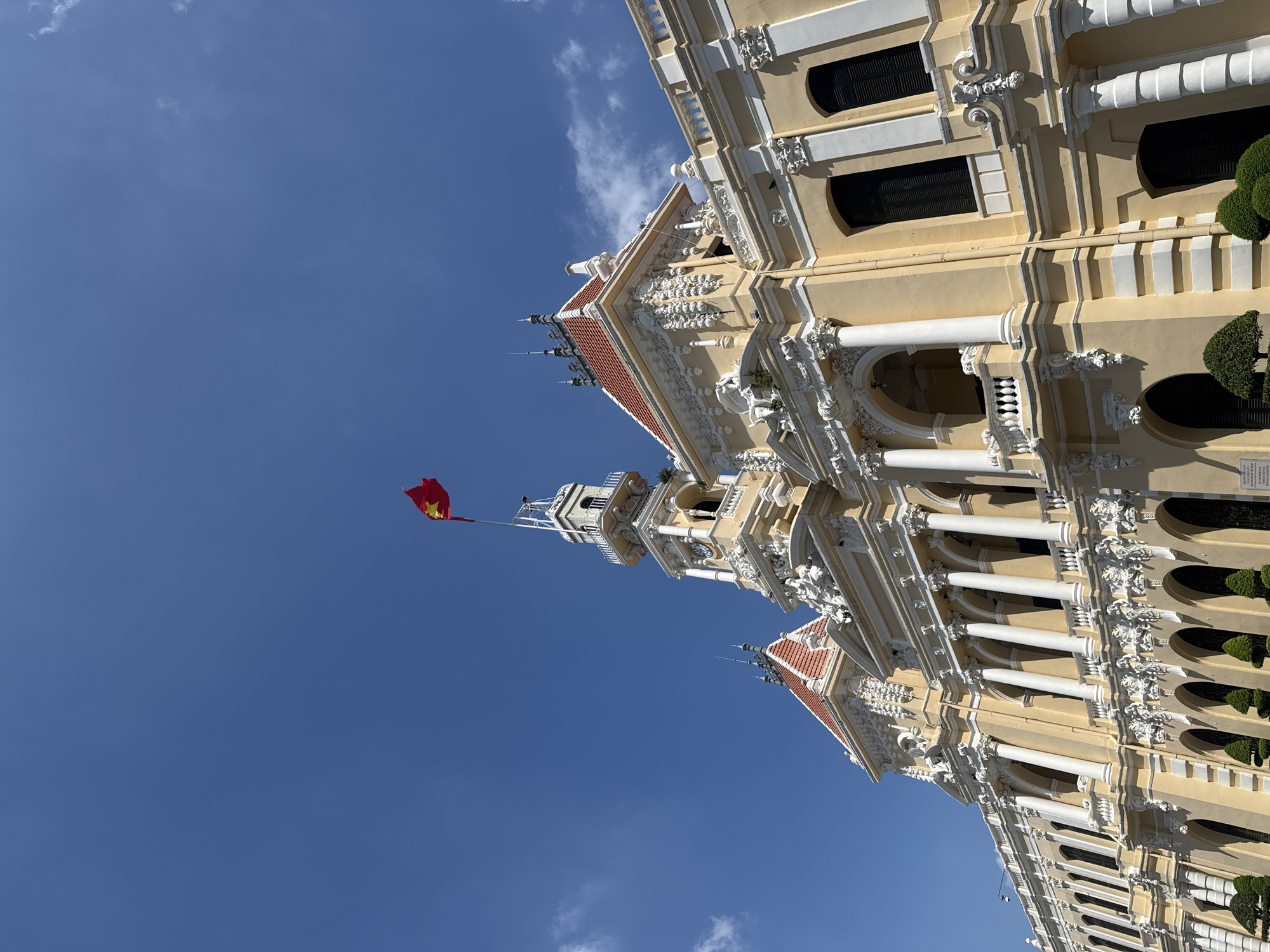 The image shows the Ho Chi Minh City Hall, a prominent French colonial-era building, located in the central district of Ho Chi Minh City, Vietnam. The building's facade is painted in a pale yellow color and features ornate white decorative elements, including columns, arches, and sculpted reliefs. A Vietnamese flag, red with a yellow star, flies from a tall flagpole atop the central tower of the building. The sky is a clear blue with a few wispy white clouds. The foreground of the image shows manicured bushes and trees, suggesting a well-maintained public space or garden area in front of the building. The architecture is grand and imposing, characteristic of colonial administrative buildings. The image is captured from a low angle, emphasizing the height and scale of the structure against the expansive sky.