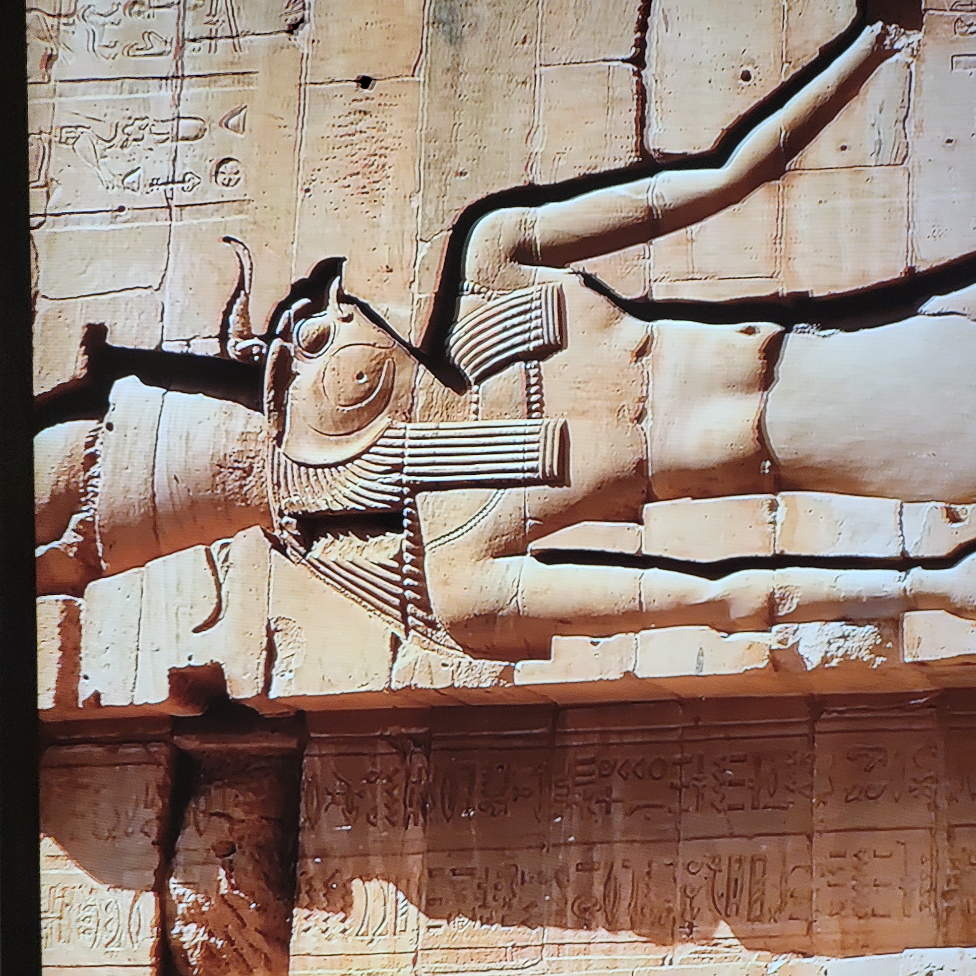 The image shows an ancient Egyptian relief carving on a wall. The carving features a horizontal figure with human-like limbs and torso, but with a bull's head and horns, and possibly a sun disk above the head. The figure wears segmented adornments around its upper body. The carving is set in a sandstone-like wall with visible blocks and rows of hieroglyphs both above and below the main figure. The lighting suggests the photograph was taken indoors or in dim sunlight. The location is not Yerevan, Armenia. This image is likely of ancient Egyptian artwork found in Egypt.