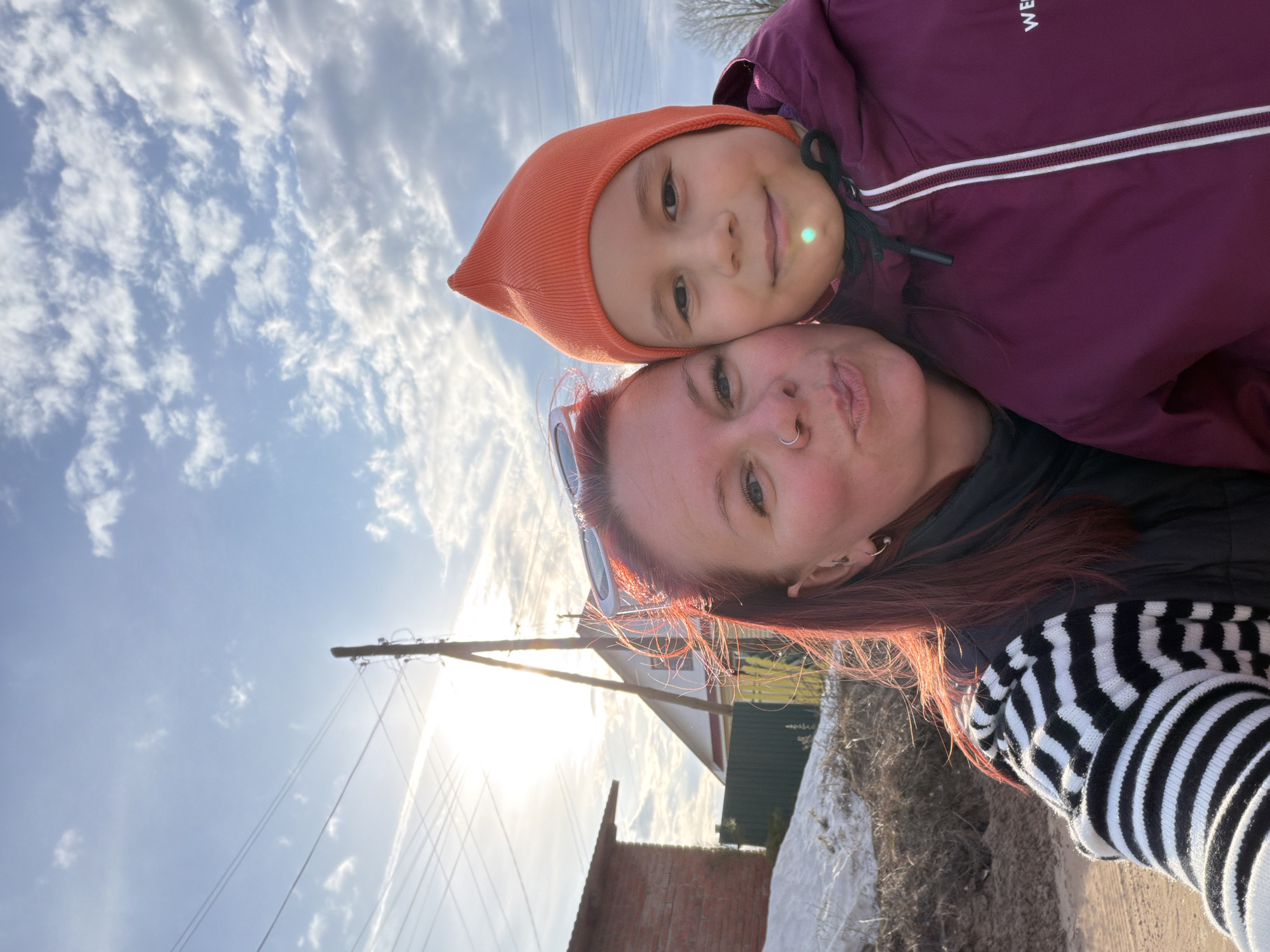 An image, rotated 90 degrees clockwise, captures a close-up selfie of an adult woman and a child outdoors in Chaikovsky, Russia. The woman, with reddish-brown hair and a nose piercing, is seen wearing a black and white striped sweater over a black jacket, with sunglasses perched on her head. She playfully makes a "kissy face" towards the camera. The child, leaning cheek-to-cheek with the woman, wears an orange knitted hat and a maroon/purple jacket, on which the text "WE" is partially visible. The child smiles gently.
The scene is set against a bright blue sky with scattered wispy clouds. The sun is shining intensely from behind the subjects, creating a strong backlight, visible glare, and a lens flare on the child's cheek. In the background, elements of a rural or semi-urban environment are discernible: a utility pole with multiple power lines, part of a white house with a green fence, and a brick structure. The ground shows dry earth, sparse vegetation, and possibly remnants of snow or ice, suggesting a late winter or early spring day. The overall mood is affectionate and joyful.