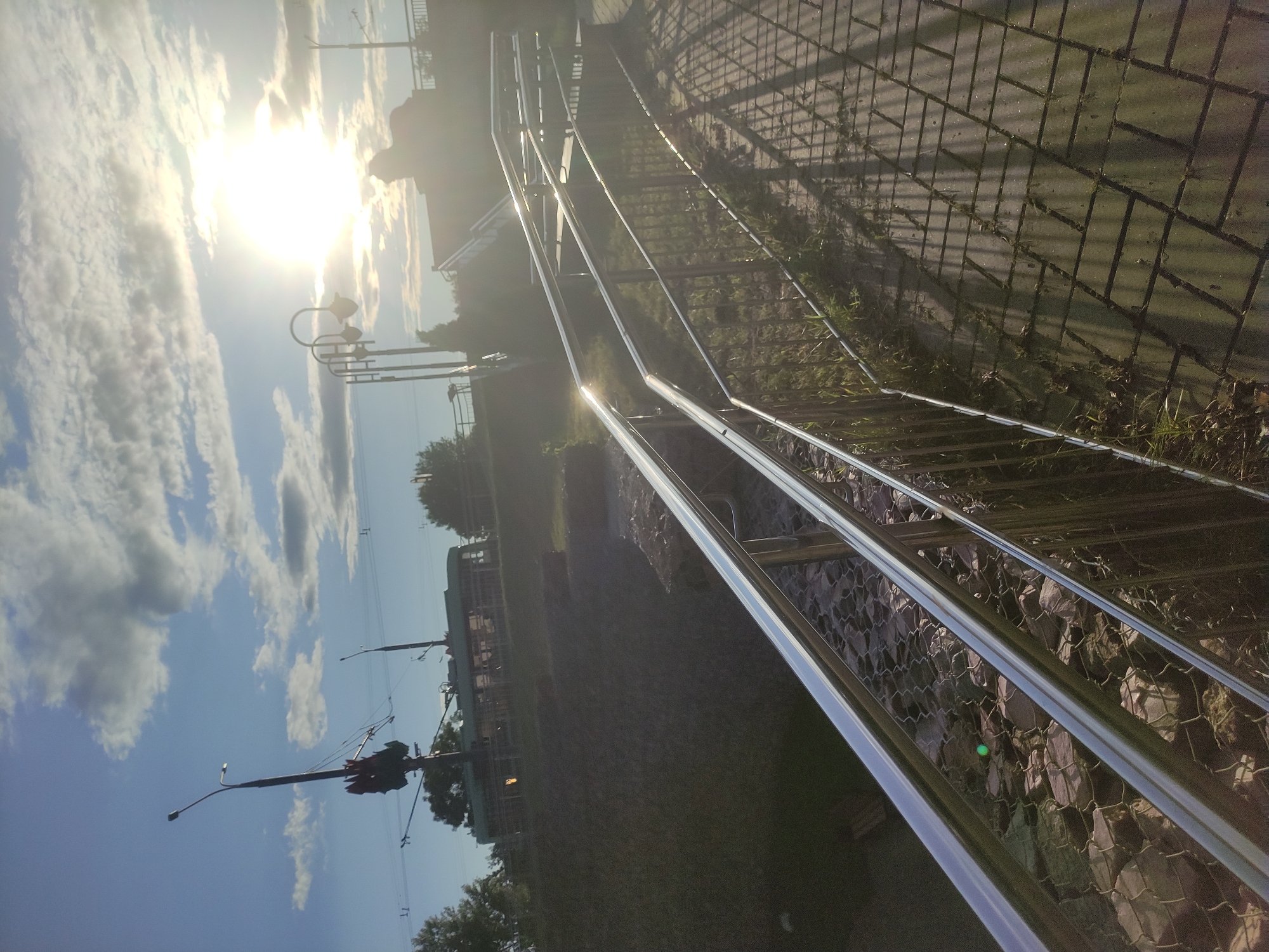 The image captures an outdoor scene in Mahilyow, Belarus, at dusk. A modern metal handrail runs diagonally across the foreground, separating a tiled pathway with shadows from a slope covered in grass and rocks, encased in wire mesh. In the middle ground, a trolleybus is visible on an elevated track, flanked by trees and streetlights. The background features a bright sun setting against a partly cloudy sky, casting a warm glow over the scene. A person is silhouetted against the sun on a distant elevated walkway. The overall impression is one of urban infrastructure blending with natural elements under a dramatic sky.