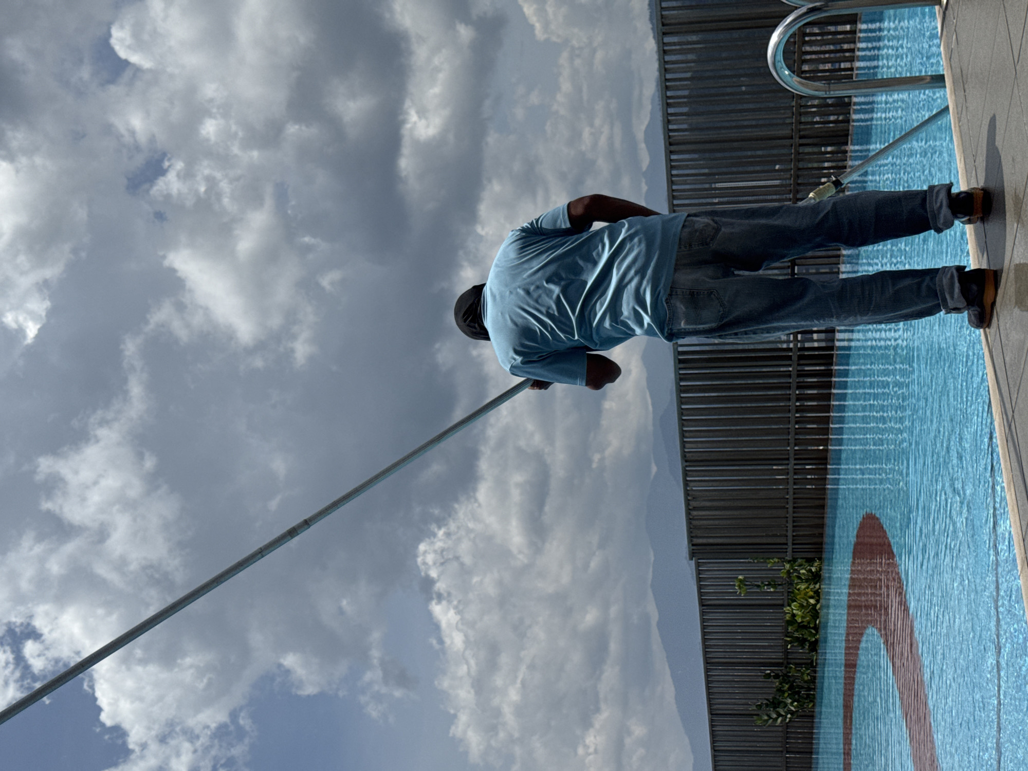 A man is standing with his back to the viewer on the edge of a swimming pool, holding a long pole. He is dressed in a light blue short-sleeved t-shirt, blue jeans, and dark shoes, with a dark cap on his head. The pole he holds extends into the light blue pool water, suggesting an activity like cleaning or maintenance.
The swimming pool has a distinctive reddish-orange arch pattern visible on its bottom. A metallic, curved handrail or ladder is partially visible at the pool's edge. Behind the pool, a dark, slatted fence or balustrade runs horizontally across the midground, with some green foliage peeking through it. In the distance, a faint outline of mountains or elevated terrain can be seen under a vast, cloudy sky.
The sky is largely covered with white and grey clouds, indicating an overcast or partly cloudy day. The overall scene suggests an outdoor setting, likely an elevated location such as a rooftop pool, consistent with a city environment like Kuala Lumpur, Malaysia. The time of day is daytime, illuminated by natural light. No text is visible in the image.