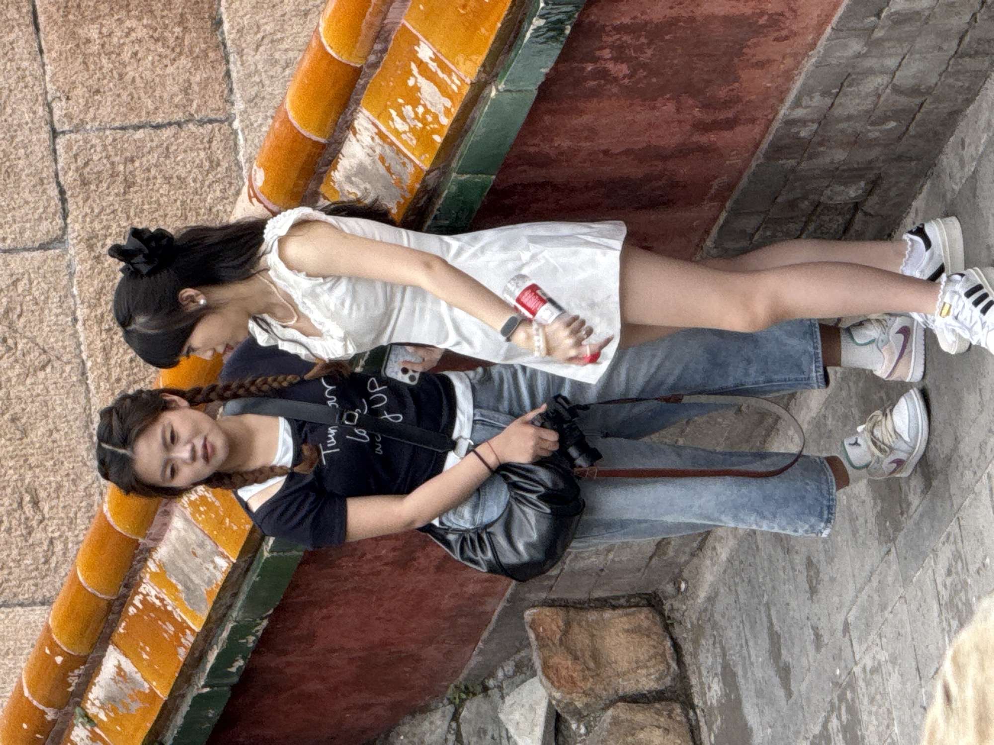 This image captures two young East Asian women standing on a paved walkway next to a traditional Chinese architectural structure, likely within the Summer Palace in the Haidian District of Beijing, China.
The woman on the left, with dark hair styled in two long braids, wears a black short-sleeved t-shirt with white text, a white undershirt, light blue cropped wide-leg jeans, and light grey and white Nike sneakers with light green accents. She has a black hobo-style shoulder bag and is holding a black camera with a lens in her right hand, while a white smartphone (likely an iPhone) is partially visible in her left hand. She is looking towards the woman on her right.
The woman on the right, with long dark hair styled with a black bow and side bangs, wears a white short-sleeved mini-dress with ruffled details, white socks with ruffled cuffs, and white and black Adidas sneakers. She wears a pearl necklace, pearl earrings, a black smartwatch on her left wrist, and multiple light-colored bracelets. In her right hand, she holds a red and white beverage can or bottle, resembling a Coca-Cola product. She is looking towards the woman on her left, leaning slightly against the wall.
The environment features a grey paved ground with rectangular blocks. Behind the women is a reddish-brown wall, possibly painted brick or stone, topped with a green-trimmed ledge and bright orange glazed roof tiles, characteristic of imperial Chinese architecture. Some tiles show signs of weathering and discoloration. Further behind and above the tiled structure is a textured, light brown stone wall. The lighting suggests an overcast day.