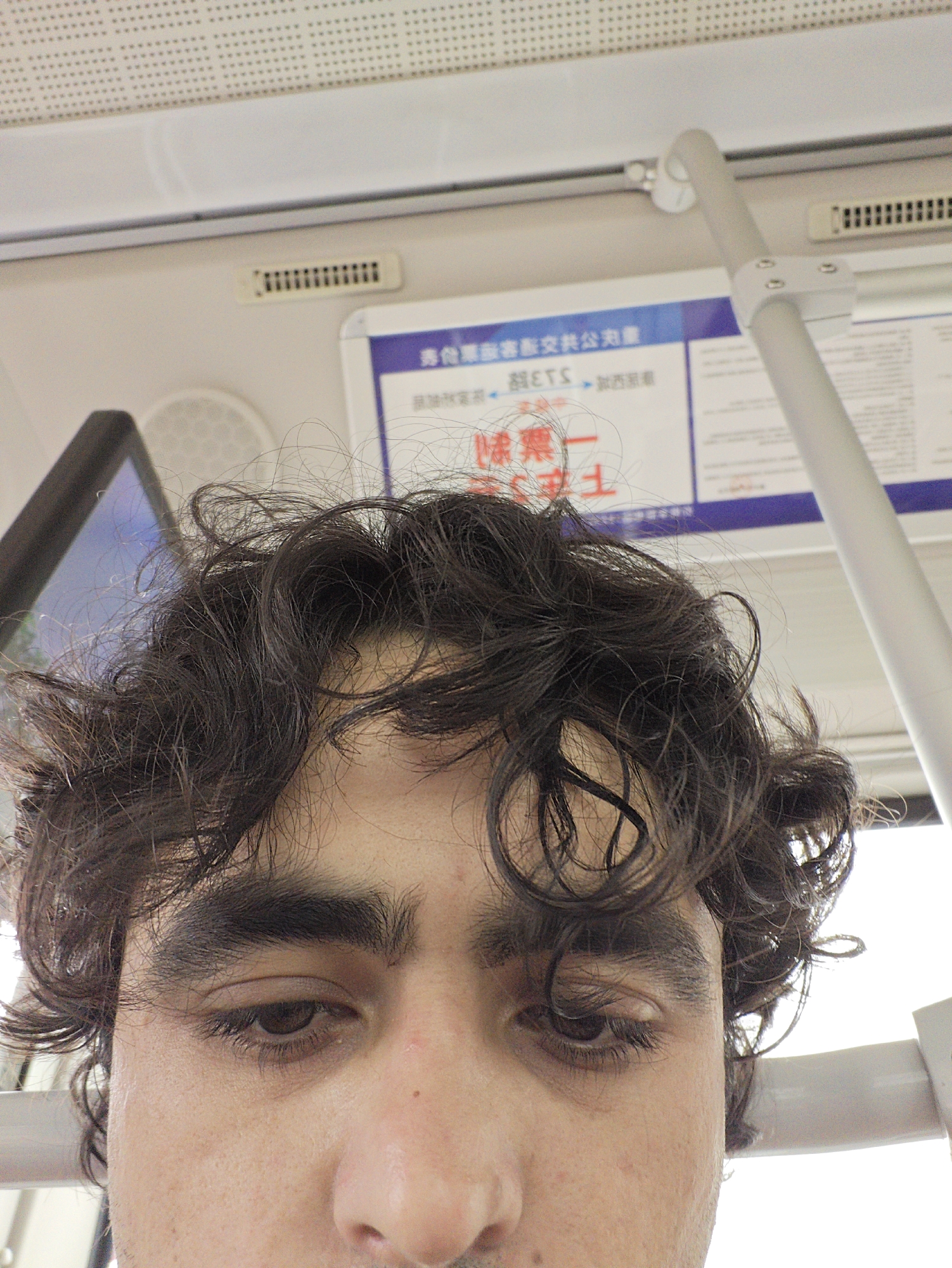 A close-up, slightly downward-angled selfie of a person with dark, curly hair, dark eyebrows, and brown eyes, appears to be taken indoors, possibly on public transportation. The person is not smiling. Above the person, in the background, is a white ceiling with small, patterned perforations. A sign with Chinese writing and a blue border is visible, as is a vertical metal pole, potentially a handrail. A dark, angled object is partially visible on the left, possibly a window or screen. The setting may be inside a bus or train based on the presence of the sign and handrail, and based on the provided location as Chongqing High-tech Zone, China.
