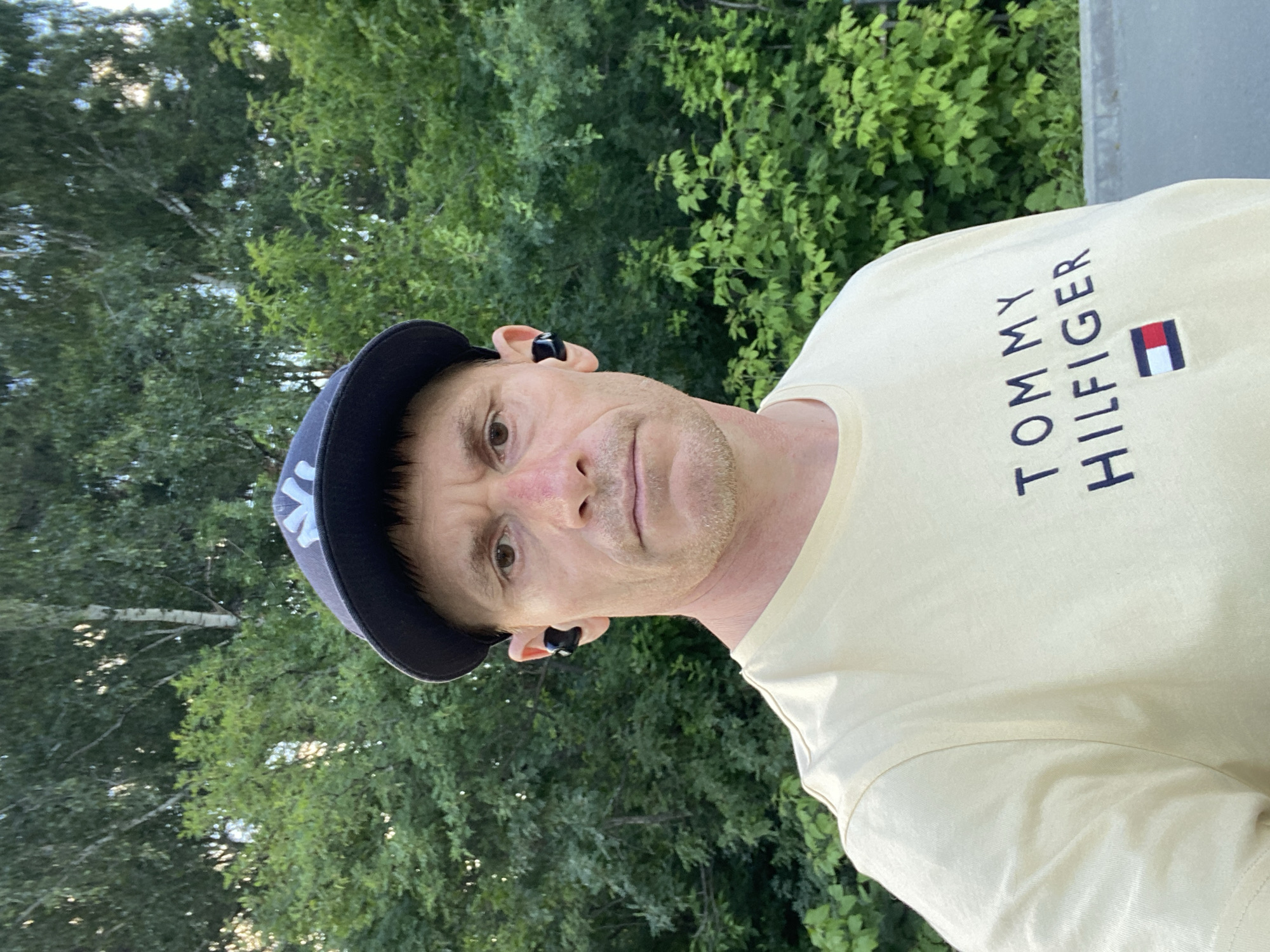 The media file is a portrait of a man taken outdoors. The man is wearing a dark blue baseball cap with a white logo and a light yellow t-shirt with the words "TOMMY HILFIGER" and a small flag logo printed on it. He also appears to be wearing black wireless earbuds. The background consists of lush green trees and foliage, suggesting a natural setting. The lighting indicates it is daytime, possibly late afternoon or early evening, with some dappled sunlight filtering through the trees. The man's expression is neutral. The image appears to be a selfie, given the angle and framing. The provided location context is Yekaterinburg, Russia.