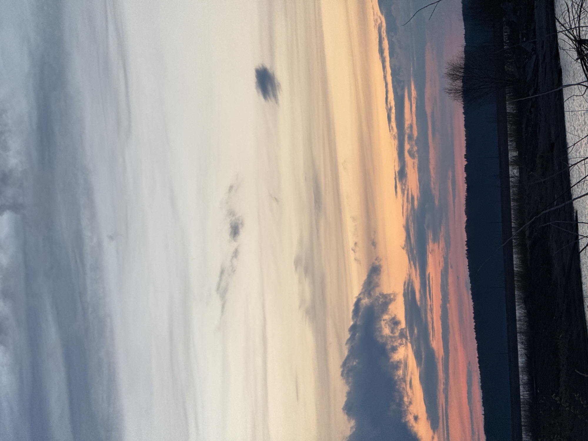 The image depicts a serene outdoor landscape at either sunrise or sunset. The sky dominates the scene, showcasing a beautiful gradient of colors. On the left, the sky is a soft blend of light blue and white with subtle streaks of light gray clouds. Moving towards the right, the colors transition to pale yellow, then warm orange, and finally deeper pinks and purples near the horizon.
Various cloud formations are scattered across the sky. There are numerous wispy, elongated clouds stretching horizontally. A more distinct, darker, and somewhat isolated cloud patch is visible in the middle-left of the sky. Further to the right, closer to the horizon, are denser, darker cloud masses that catch the warm hues of the setting or rising sun.
The horizon line runs horizontally across the bottom section of the image, formed by a dark, silhouetted landmass, likely a distant tree line or hills. To the far right, a body of water or a reflective shore is visible, with dark, bare branches extending into the frame from the right edge. The general mood conveyed is peaceful and tranquil. There is no visible text in the image.