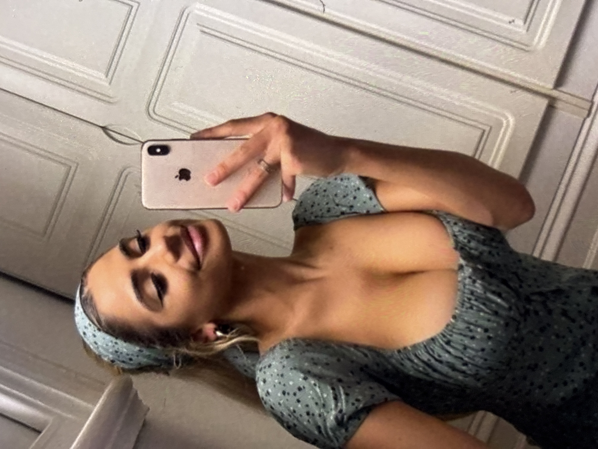 In a close-up, waist-up selfie, a young woman with tan skin and long blonde hair tied back with a light blue patterned bandana poses in front of a white paneled door. She wears a short-sleeved, green polka dot dress with a low-cut neckline. Her left hand holds a rose gold iPhone, angled to capture her reflection. She has on hoop earrings and a ring on her left ring finger. Her right arm is bent and rests on her hip. The background appears to be an interior setting, possibly a bathroom or bedroom, with the white paneled door and trim of a door frame visible.