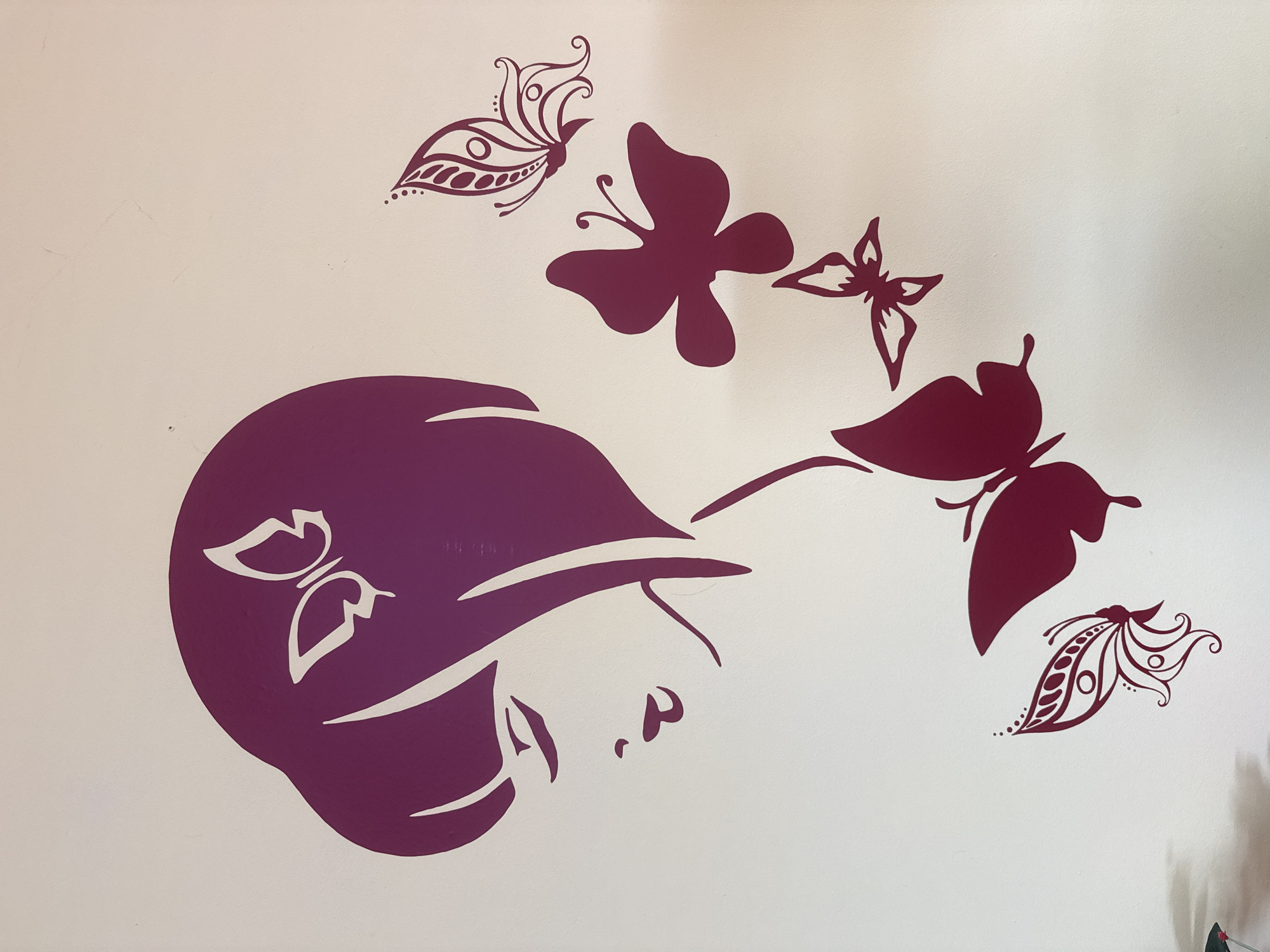 The image shows a decorative wall with purple decals. The decals depict a stylized silhouette of a woman's face in profile, facing right, with butterflies around her. The silhouette includes a butterfly design on the hair. The butterflies are in different shapes and styles, with varying levels of detail. The wall appears to be white. This image was taken in the city of Pleven, Bulgaria.