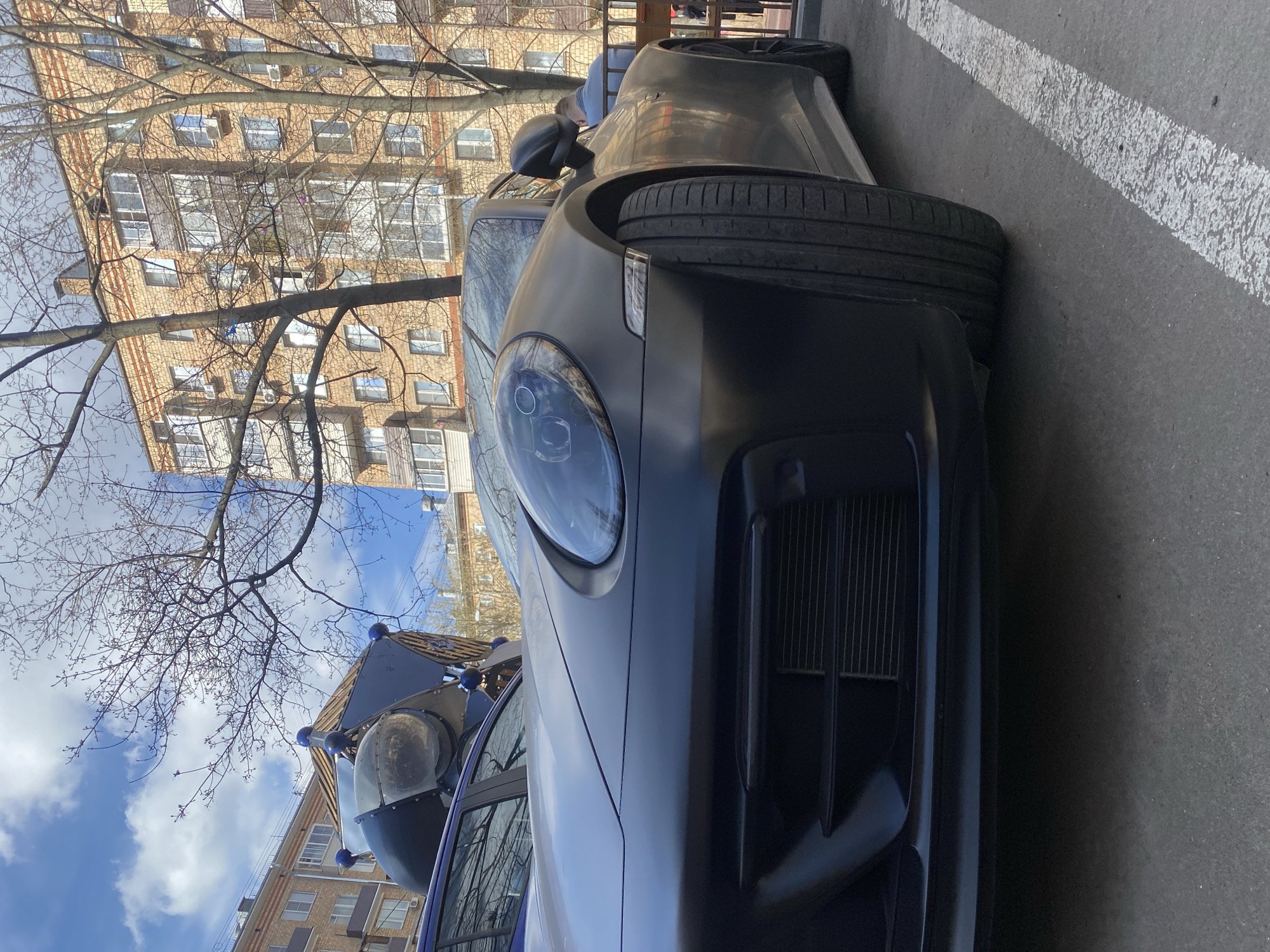 The image presents a low-angle, close-up view of a dark-colored Porsche parked on an asphalt surface with a white parking line. The front of the car is prominently displayed, showcasing its headlight and grille. To the left, a children's playground structure with blue accents is visible. The backdrop features a multi-story brick building with multiple windows, typical of urban architecture. Above the building and playground, the sky is visible with sparse tree branches in the foreground, possibly suggesting early spring or late autumn. Given the architectural style of the building and the overall urban setting, it is plausible that the location is Moscow, Russia.
