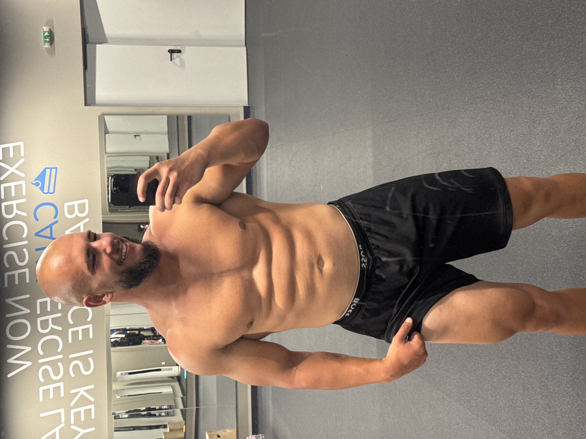A shirtless man with a shaved head and beard is taking a mirror selfie in a gym. He is wearing black athletic shorts. The gym has a gray floor and mirrors on the wall with motivational text written in white and blue. The text on the wall appears to be in English and partially reads "EXERCISE IS KEY" and "WORK NOW". There are also some gym equipment and doors visible in the background. The overall lighting suggests an indoor setting during the day. The man's physique is muscular, with visible abs. He is smiling at the camera. The location is specified as Sofia, Bulgaria.