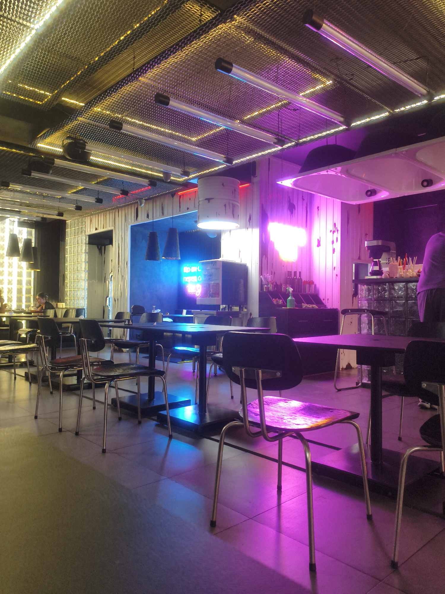 This image depicts the interior of a modern cafe or restaurant with a vibrant, contemporary design. The space is characterized by its use of dramatic lighting, primarily neon and LED, casting colorful hues of pink, purple, and blue across the scene. The ceiling features a metallic, patterned mesh with integrated yellow LED lighting, creating an industrial yet chic aesthetic. Several fluorescent tube lights are also suspended from the ceiling.
The furniture consists of dark-colored tables and chairs with minimalist metal frames. Some chairs have wooden seats and backs, while others appear to be fully upholstered in dark fabric. The flooring is tiled, reflecting the colorful lighting.
In the background, a neon sign glows in bright blue against a textured wall, adding to the overall dynamic atmosphere. There are also elements of decor such as bottles on shelves and a coffee machine visible. A person can be seen seated at a table in the far left of the image, appearing to be engaged with something on their table. Another person, dressed in a purple shirt and dark shorts, stands near the counter area on the right side of the image. The environment suggests a lively and stylish urban establishment.
The location is identified as a cafe in Kremenchuk, Ukraine.