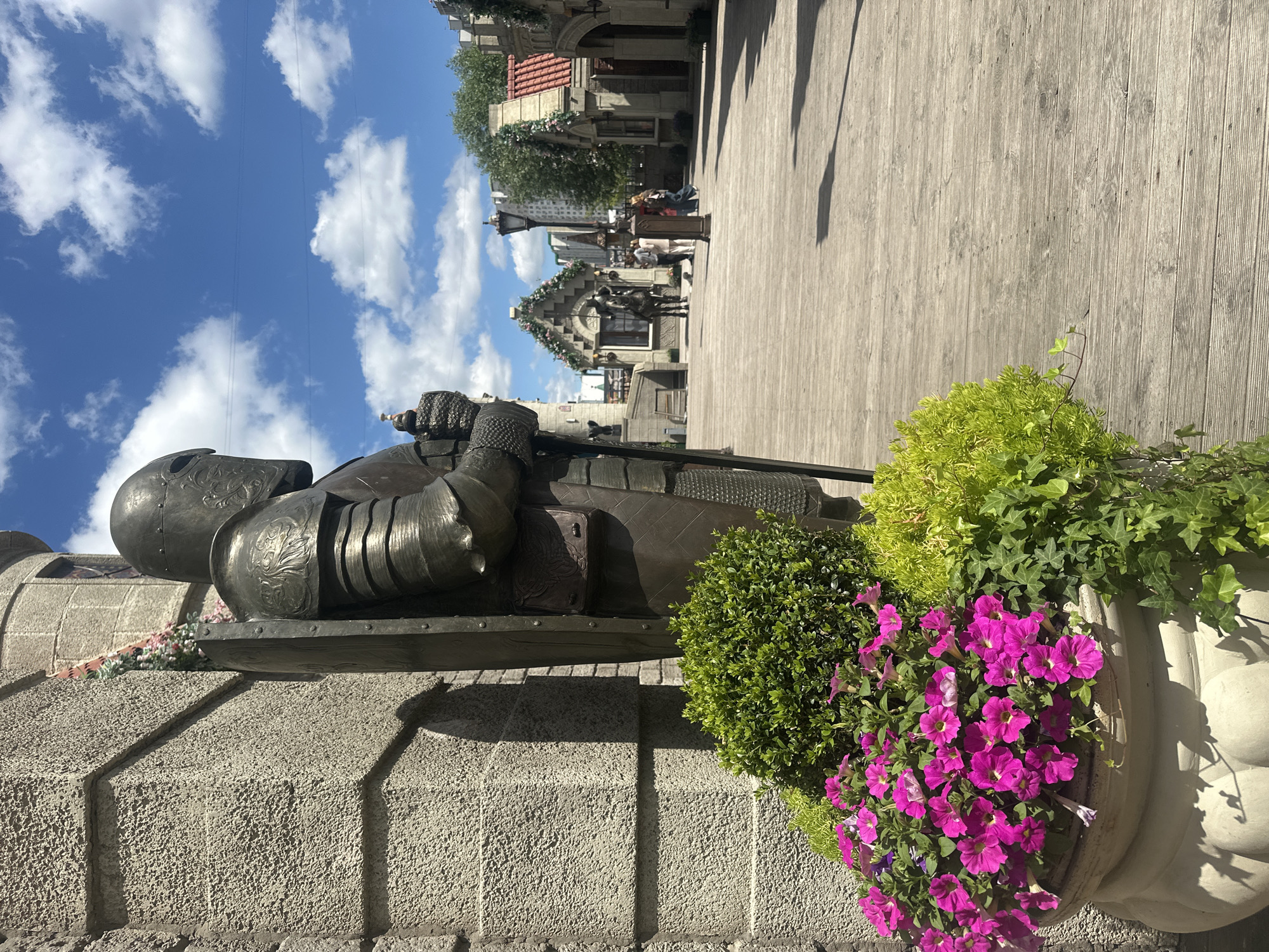 The image shows a bronze statue of a knight in full armor, standing next to a stone wall and a large planter filled with vibrant pink petunias and lush green foliage. The statue is depicted in a protective stance, with a shield held before it. In the background, a charming street scene unfolds, featuring buildings with red-tiled roofs, trees, and more greenery. Several people can be seen in the distance, suggesting a public space or a pedestrian area. The scene is bathed in bright sunlight, casting distinct shadows, indicating it's daytime, likely late morning or early afternoon. The sky is a clear blue, dotted with fluffy white clouds. The wooden boardwalk or pavement in the foreground suggests an outdoor plaza or promenade. The architecture and the presence of a knight statue might hint at historical or themed areas within Moscow, Russia. There is no visible text in the image. The overall mood is bright and cheerful, with a touch of historical grandeur evoked by the knight statue.
