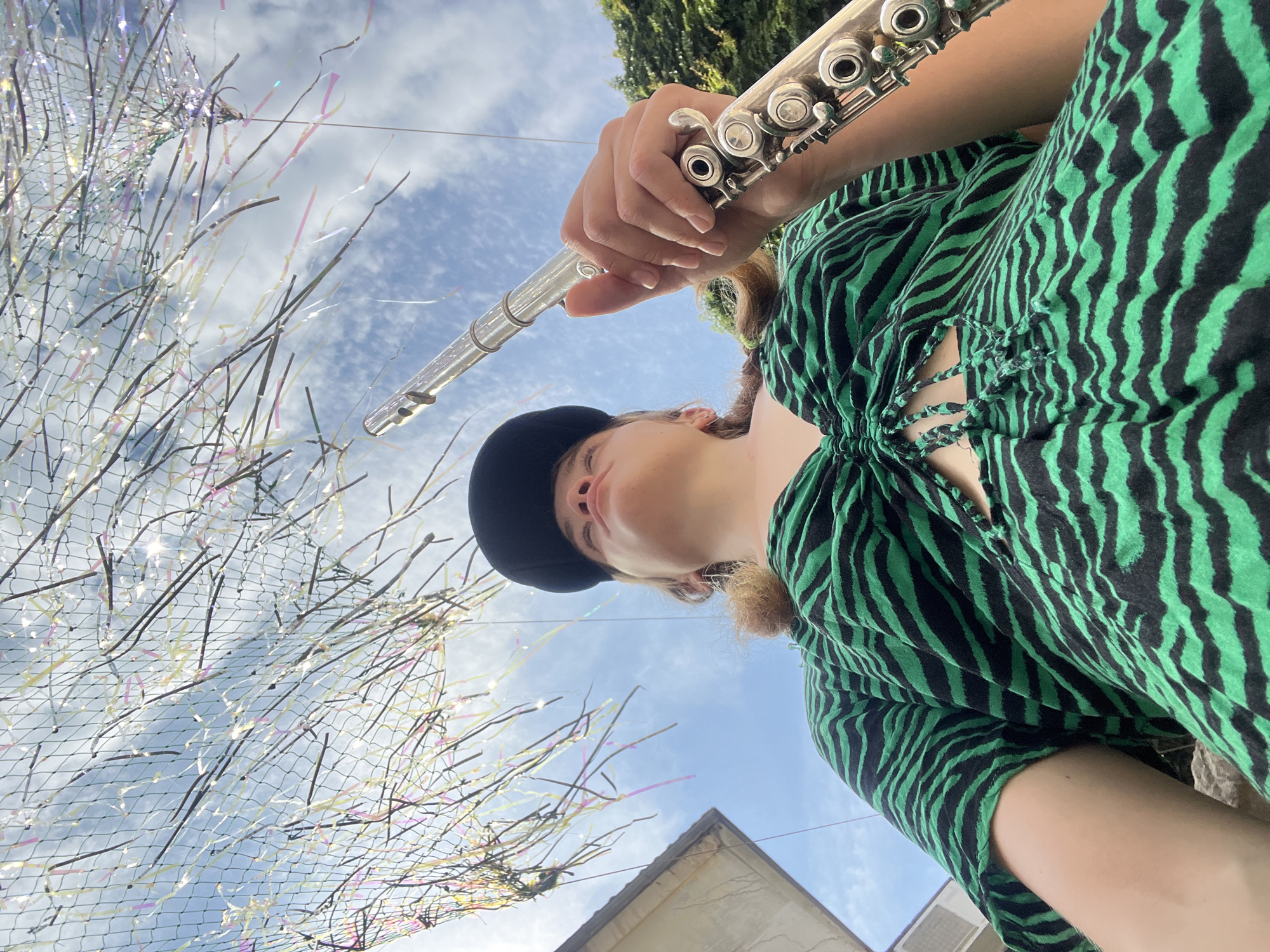 A young person wearing a black baseball cap and a green and black striped top is holding a silver flute up to the sky. They are looking upwards at a net strung with iridescent streamers. The sky is blue with scattered clouds. The scene is shot from a low angle, looking up at the person and the sky. In the background, a portion of a building and some green trees are visible. The location is the city of Budva, Montenegro.