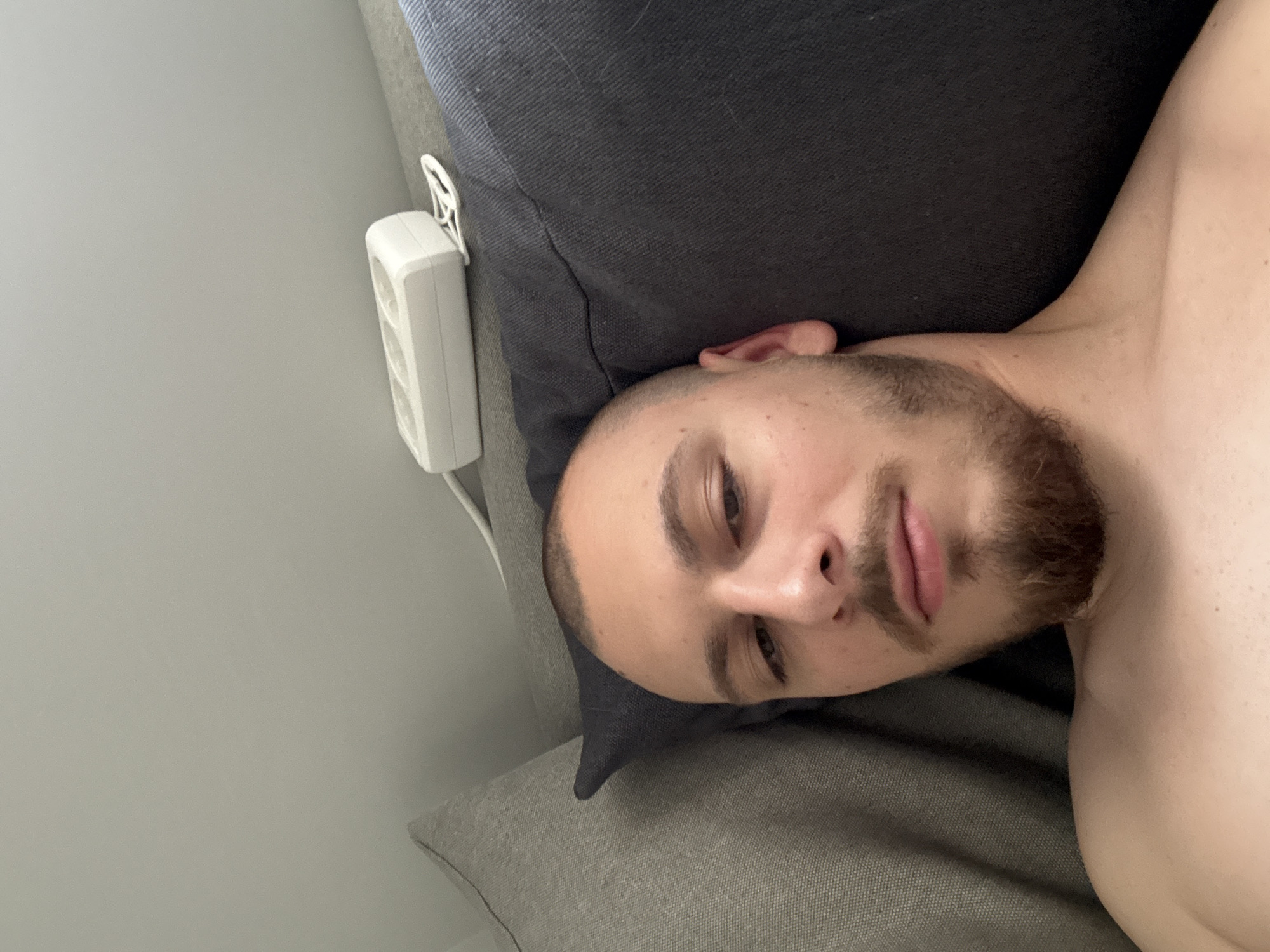 A close-up, slightly angled photograph depicts a man lying on a gray couch. His head rests on a darker gray pillow. He has short hair and a beard. His upper body is visible, appearing shirtless. To the left of the pillow, a white electrical outlet strip is mounted on the wall. The background consists of a light gray wall. The location of the photo in the city of Stara Zagora, Bulgaria is not evident in the photograph.
