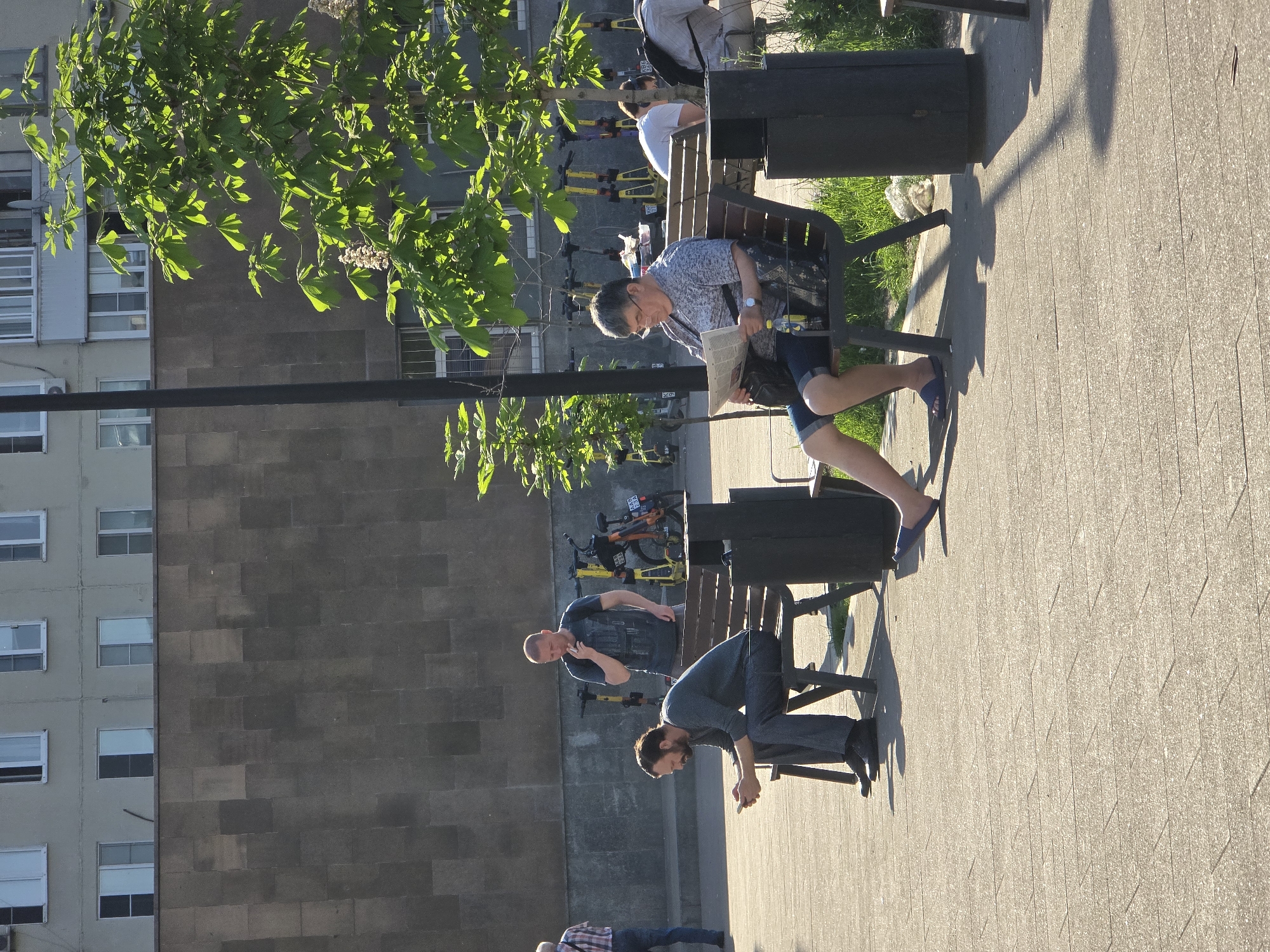 The image depicts a sunny day in what appears to be a park or public area in Moscow, Russia, judging by the architecture and the presence of shared mobility scooters (presumably Yandex scooters). A man is seated on a park bench, reading a newspaper. Another man stands behind him and to the left and is talking on a phone. A third man has his hands clasped on a different part of the bench. In the background, several shared mobility scooters are lined up. A tree provides partial shade over the bench and the people. A multi-story building is visible to the left of the scene. The ground is paved with interlocking concrete pavers. There are trash cans alongside the park benches.