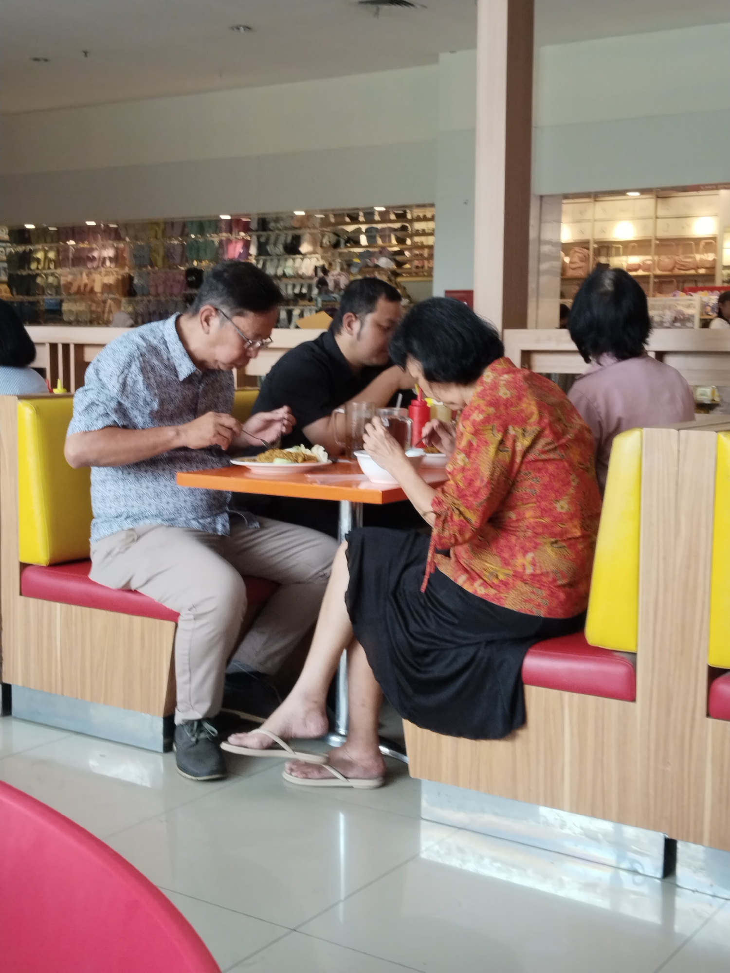 In a casual dining establishment, possibly within a mall or food court setting in Kudus, Indonesia, four individuals are seated at an orange-topped table. The man on the left, wearing a blue patterned shirt and light-colored trousers, is engaged in eating. A woman to his right, dressed in a vibrant red and yellow patterned top and a black skirt, is also eating. Behind her, another woman in a light pink top is partially visible, and to the left of the man, a man in a black shirt is seated. The table is set with plates of food and drinks. The seating consists of red and yellow benches within a wooden booth structure. The background reveals shelves stocked with various items, likely merchandise, and part of the interior architecture of the building, including columns and lighting.