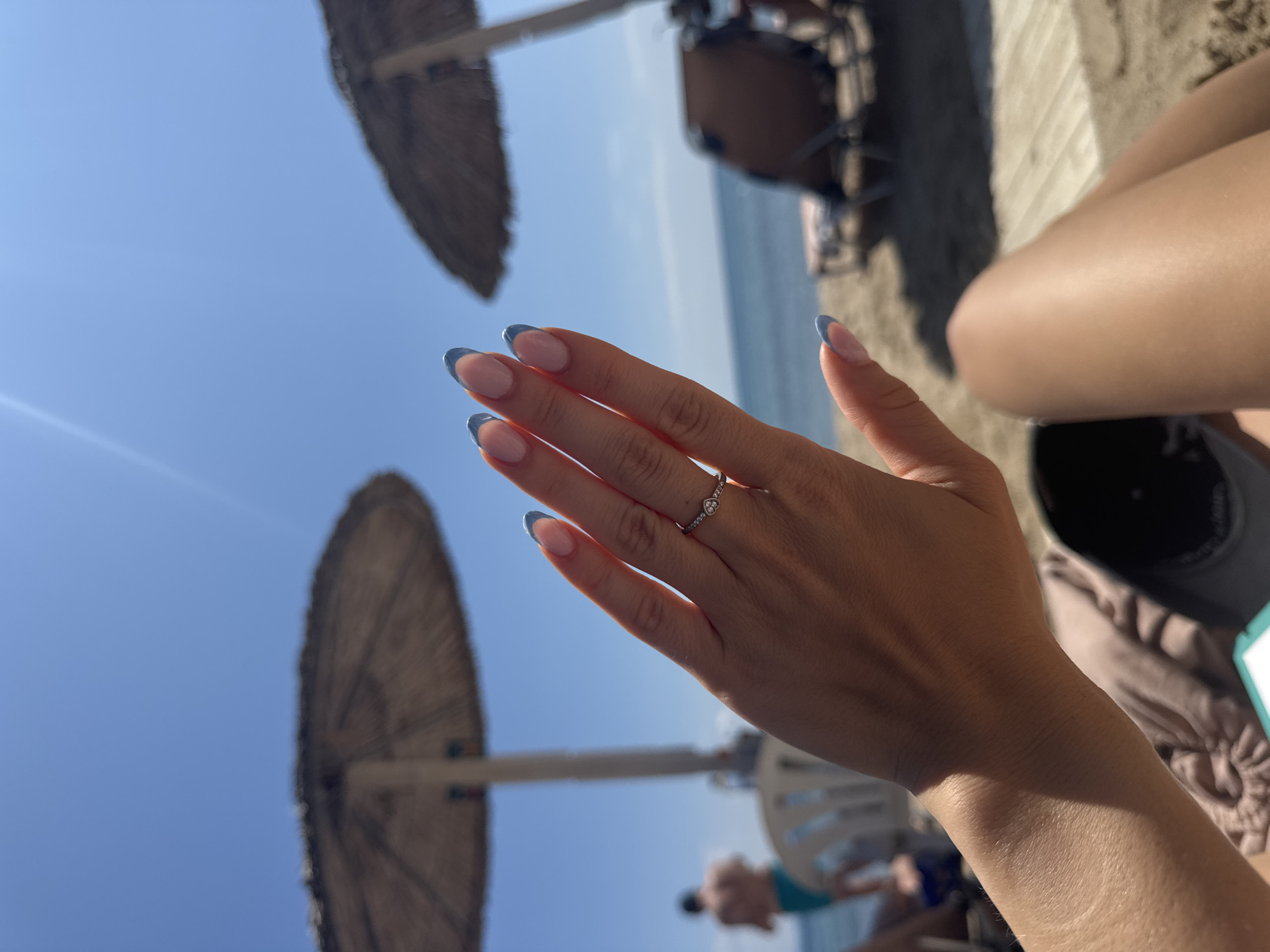 A first-person perspective shot shows a woman's hand with light blue French manicure and a diamond ring with a flower design, fingers extended upwards against a bright blue sky. The hand is positioned in the foreground, slightly to the left of the frame. In the background, two thatched umbrellas provide shade over beach chairs. A man in teal swimming trunks is visible in the blurred distance near the water's edge. The setting appears to be a beach, likely in Stavros, Greece, given the context. The overall scene conveys a relaxed, sunny atmosphere.