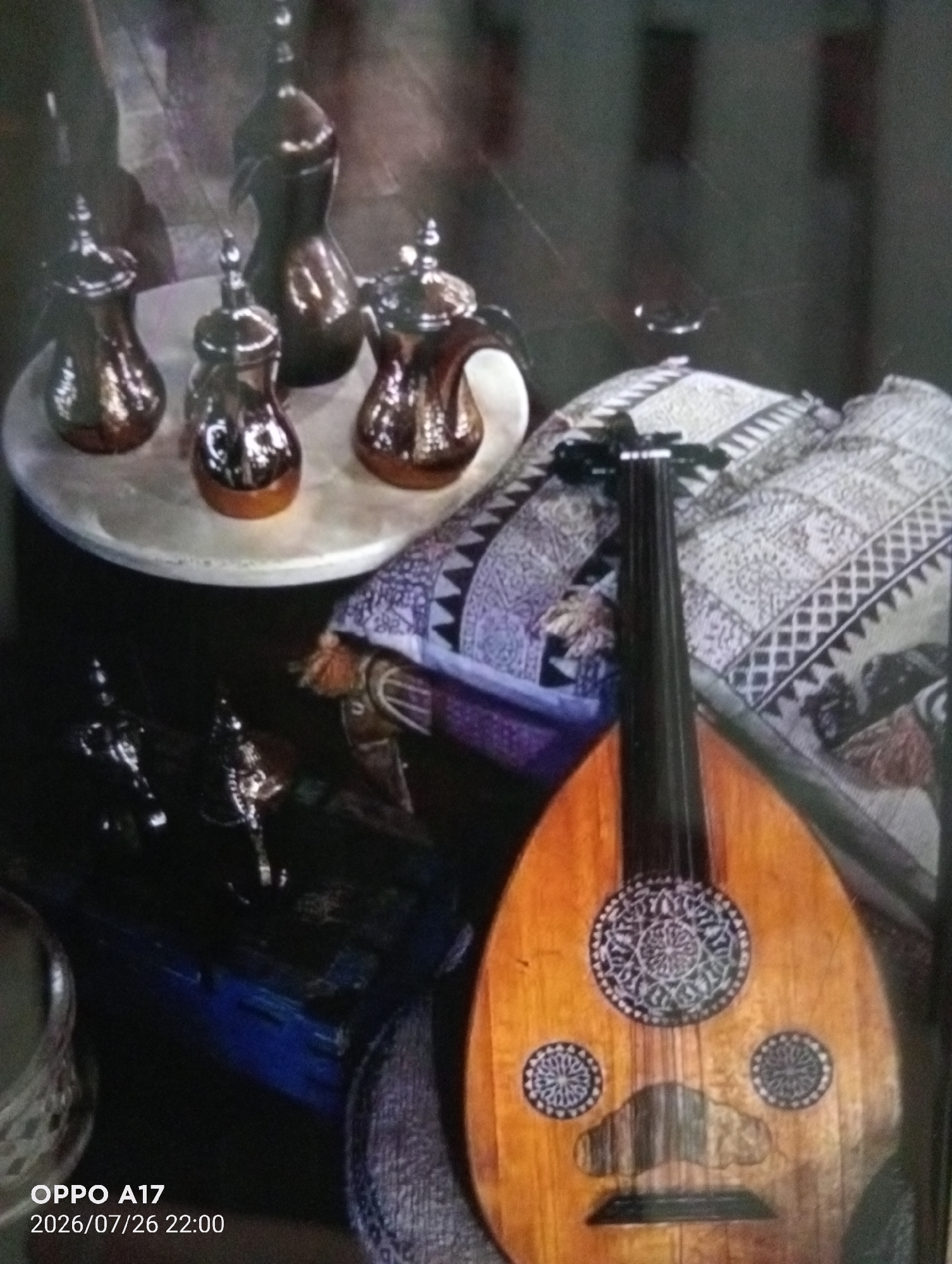 The image depicts a still life arrangement featuring traditional Arabic coffee pots, a musical instrument resembling an oud, and decorative items. Four gleaming, metallic coffee pots with ornate handles and spouts are placed on a round, light-colored surface. To the right, a dark-colored musical instrument, likely an oud, with a rounded body and intricate rosettes on its soundboard rests against a patterned textile. The background is dimly lit and appears to be an indoor setting, possibly a home or shop. The lower left corner of the image displays a timestamp reading "OPPO A17 2026/07/26 22:00". There are no people or discernible actions taking place. The scene does not contain any specific visual indicators that would allow for the precise determination of its location within the city of Құмөзек, Kazakhstan.