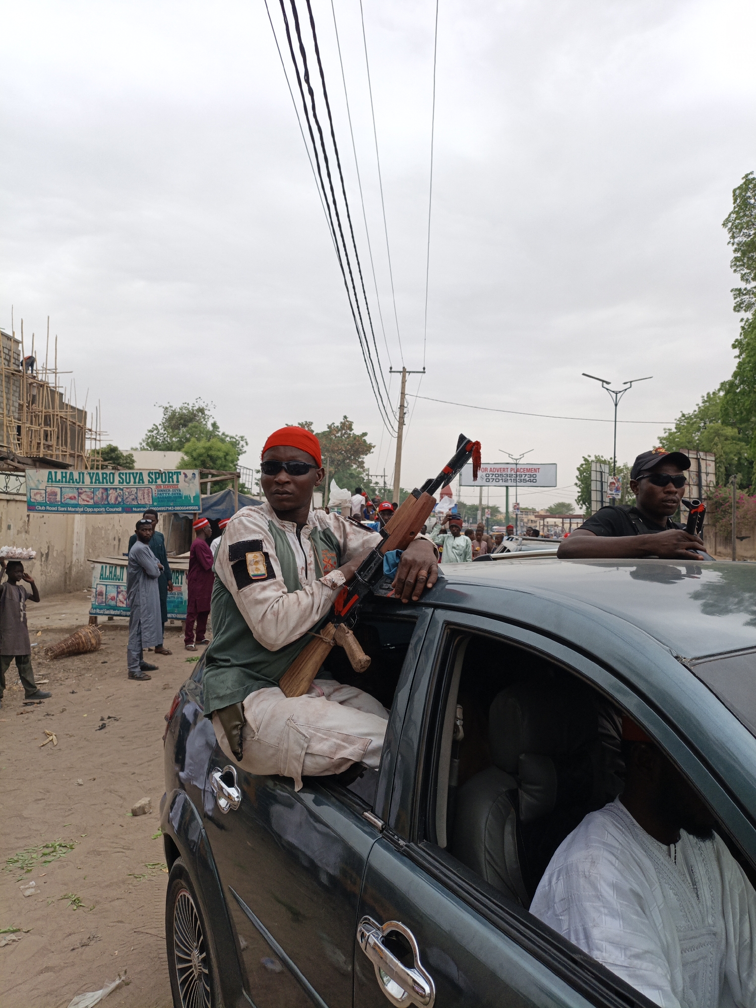 The image shows two men in a dark teal sedan. The man in the passenger seat is wearing a red head covering, sunglasses, and an olive green overshirt. He is holding an automatic rifle. The man in the back of the car is wearing a white robe. The man standing by the roof of the car is wearing a black shirt and a black hat. They are in Giginyu, Nigeria. In the background there is a sign that reads "ALHAJI YARO SUYA SPORT". There are a number of people standing and walking around. There are electrical wires running across the top of the frame. There is a building under construction in the background.