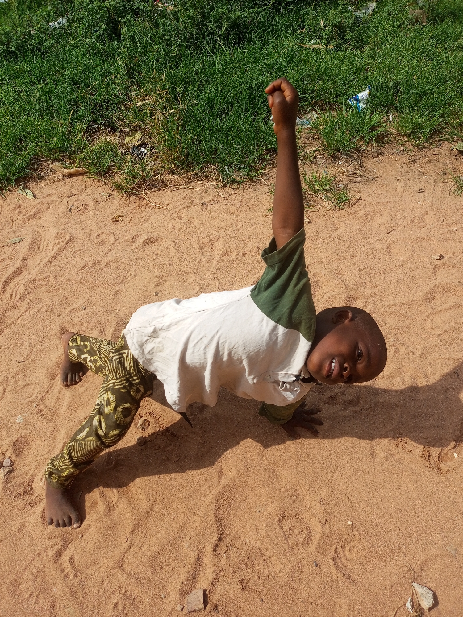 In Tempure, Nigeria, a young boy is captured in a playful moment on a sandy ground. He is positioned on his hands and knees, with his right arm raised high in the air, his fist clenched. The boy is wearing a white t-shirt with green accents on the sleeves and a pair of patterned green pants. His gaze is directed towards the camera with a wide smile, revealing his teeth. The ground beneath him is sandy and appears to be in an outdoor setting, with a patch of green grass and some scattered debris visible in the background. The lighting suggests it is daytime, with shadows indicating the sun's position. The scene is informal and captures a candid expression of joy and energy from the child.