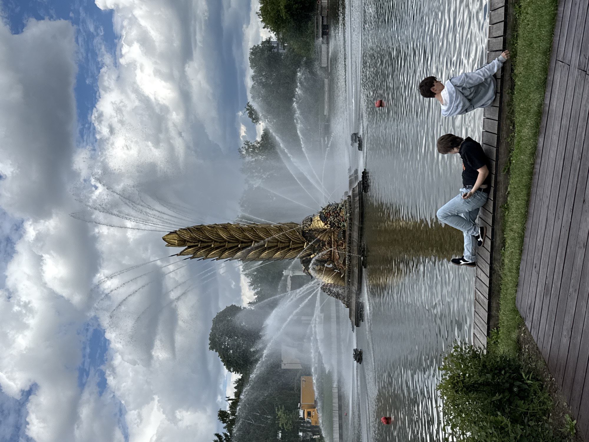 The image depicts the "Friendship of Nations" fountain in Moscow, Russia. Two individuals are seated on a wooden platform facing a large body of water. The fountain, a prominent golden structure resembling a sheaf of wheat, is active, with water jets spraying upwards and outwards. The sky is partly cloudy with dramatic cloud formations. Trees and other buildings are visible in the background. One person is lying down, looking at their phone, while the other is sitting up, looking out at the water. A yellow truck is parked on the far left of the image. The overall scene suggests a public park or recreational area in Moscow.