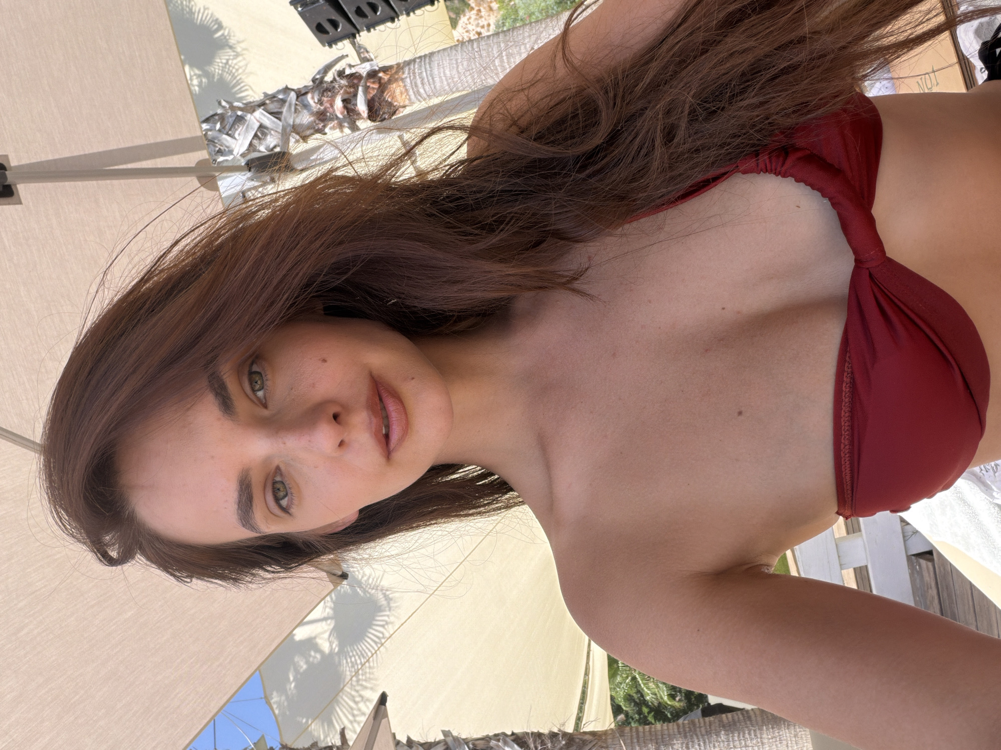 A young woman with long, brown hair and green eyes, wearing a red bikini top, poses for a selfie under a white umbrella. She is outdoors, with palm trees and a glimpse of the sky in the background. The image appears to be taken during the daytime, likely in a tropical or Mediterranean setting.