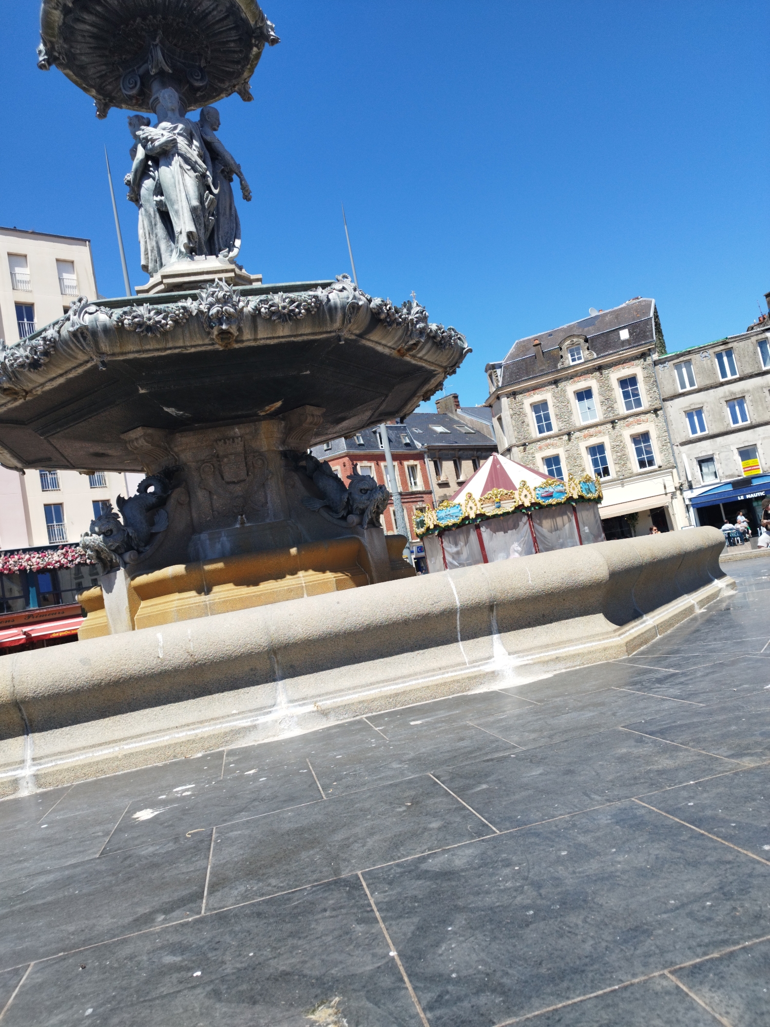 The image shows a close-up view of a stone fountain in Cherbourg-en-Cotentin, France, with a bright blue sky overhead. The fountain features intricate carvings and statues. In the background, there are several buildings, including a carousel with red and white stripes and decorative panels depicting boats. Some people are visible in the distance near the carousel and a building with a blue awning. The ground in the foreground is paved with large, dark gray tiles.