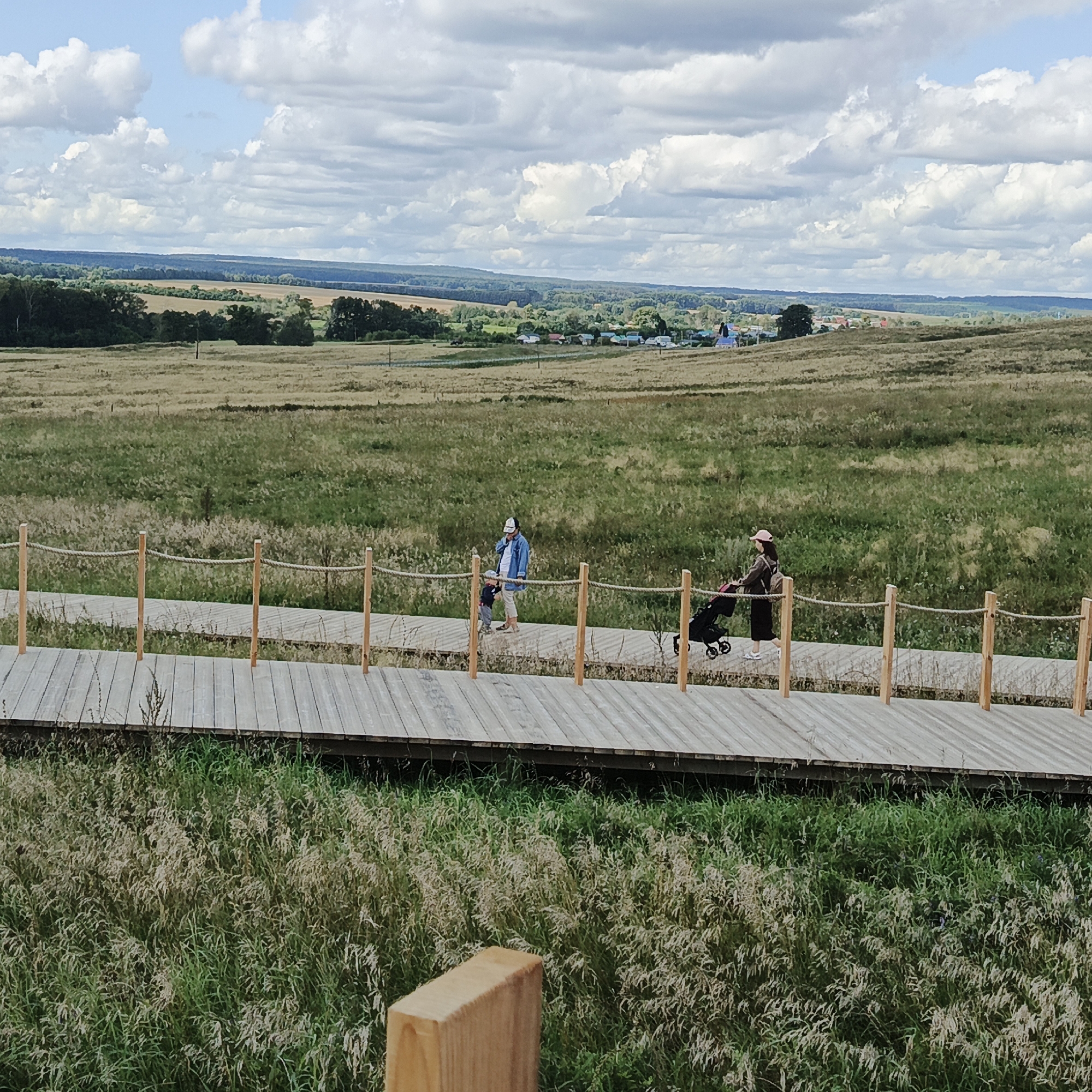 In Verkhnie Termy, Russia, a wooden boardwalk winds through a grassy field under a partly cloudy sky. Two people are walking on the boardwalk. One person, dressed in a denim jacket and white cap, is walking with a small child. Another person, wearing a dark outfit and a pink hat, is pushing a stroller. The boardwalk is bordered by wooden posts and rope railings. In the background, rolling hills and a distant village are visible. The overall scene suggests a peaceful day spent outdoors in a rural setting.