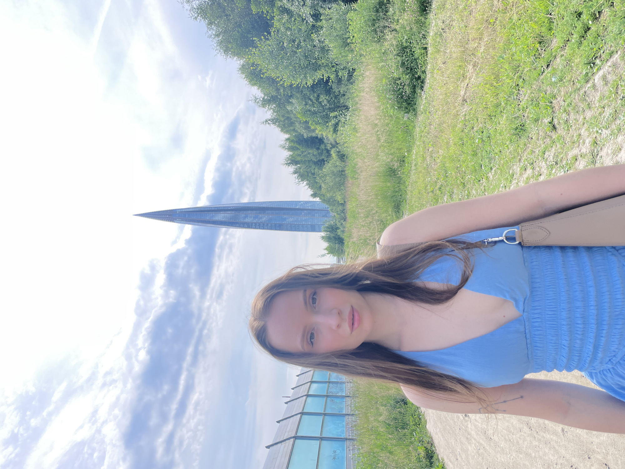 The image is a portrait of a young woman outdoors, with Saint Petersburg, Russia, as the location. She is facing the camera, looking directly at it with a neutral expression, and her head is tilted slightly. She has long, brown hair that falls around her shoulders and chest. She is wearing a light blue, ruched dress with a V-neckline and a beige purse slung over her shoulder.
In the background, there is a tall, slender, glass-fronted building that tapers to a point, characteristic of modern architecture. To the right of this building, there is a lower, modern building with a glass facade. The setting appears to be a park or open area, with lush green grass and trees bordering a dirt path. The sky is partly cloudy with some bright patches, suggesting it is daytime, likely late morning or early afternoon, under fair weather conditions. The lighting indicates a sunny day. The overall mood is relaxed and casual, as if the woman is enjoying a day out. There is no visible text in the image.