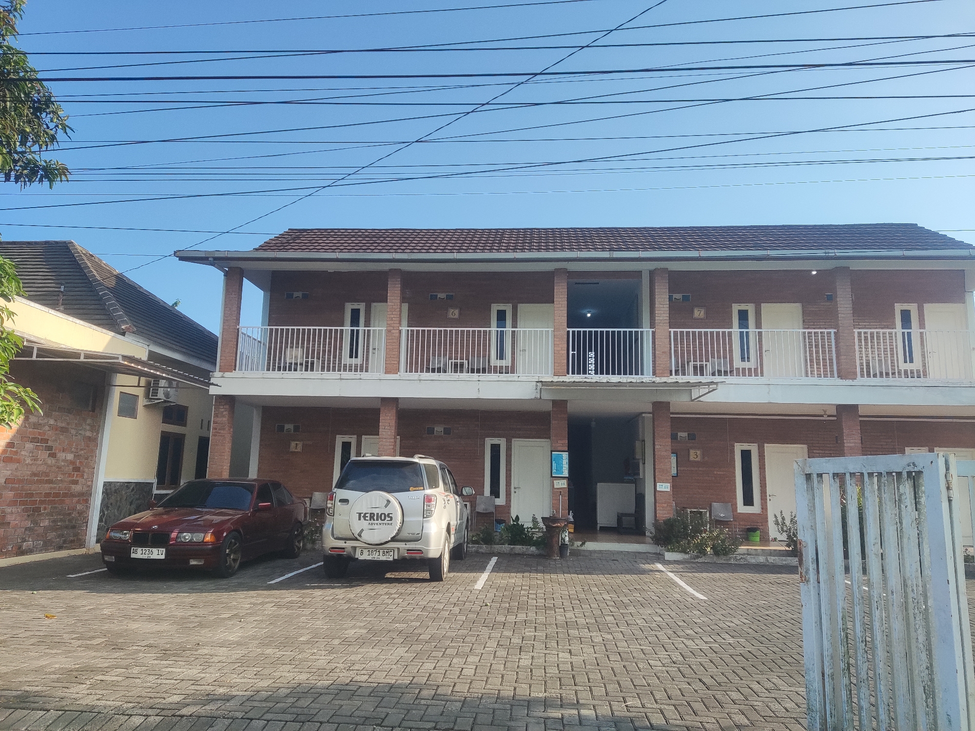The image shows a brick building with multiple units and a paved courtyard. Two cars are parked in the courtyard: a dark red BMW coupe and a silver SUV with "TERIOS ADVENTURE" written on the back. The building appears to be a guesthouse or apartment complex, with several doors and windows visible. A white railing runs along the second-floor balcony. Overhead, power lines crisscross against a clear blue sky. The scene is likely a residential or commercial area in Tlogoadi, Indonesia, given the presence of what appear to be Indonesian license plates on the vehicles. The overall impression is one of a well-maintained property on a bright, sunny day.