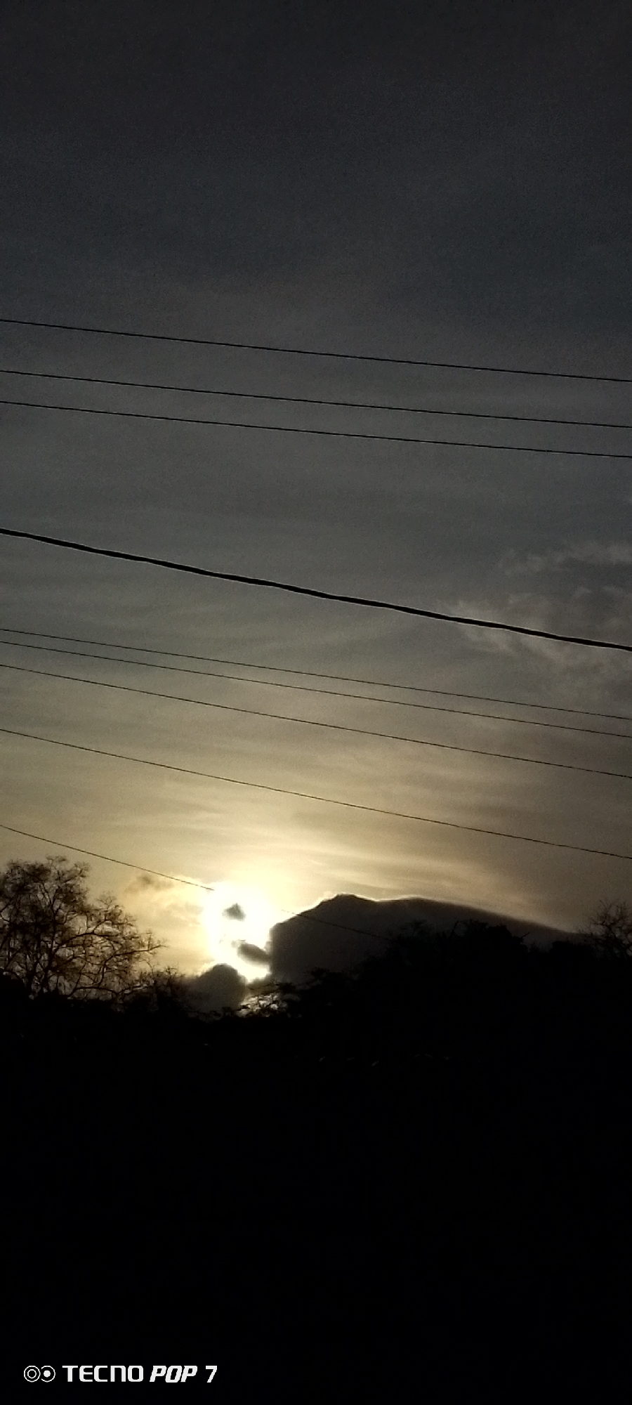 The image shows a silhouette of a mountain or large hill, partially obscured by trees and foliage at its base. The sky is overcast, with a bright patch suggesting the sun is setting or rising behind the mountain. Overhead power lines are visible against the sky. The location is likely near Altagracia, Venezuela.