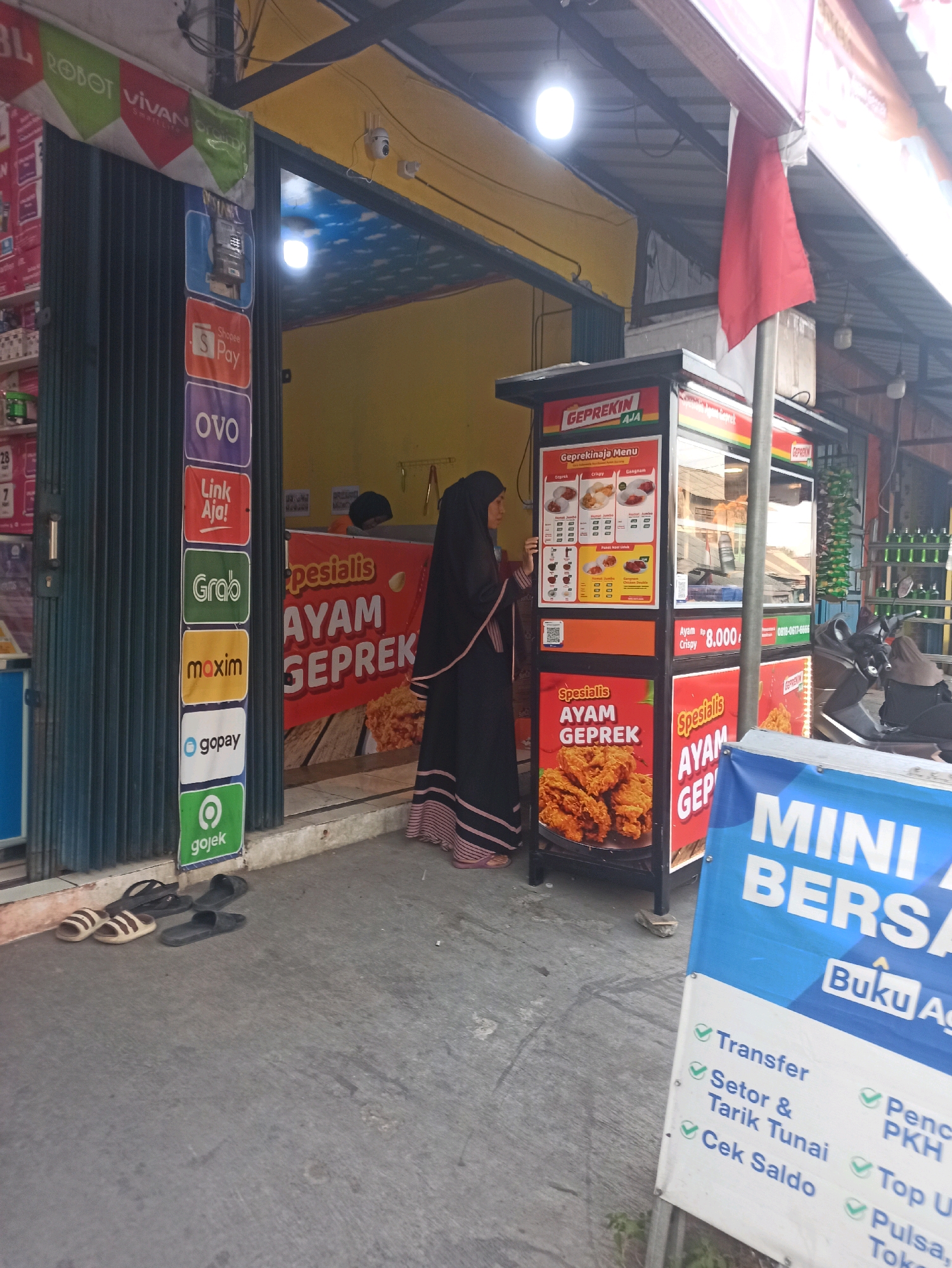 A woman wearing a black hijab and dress stands to the left of a food stall in Telagamurni, Indonesia. The stall, branded "GEPREKIN AJA," sells "Spesialis Ayam Geprek" (Specialty Fried Chicken). A menu board is visible on the stall, displaying various chicken meal options and prices. The stall is decorated with images of fried chicken and a red and white Indonesian flag. To the left of the stall, a vertical strip displays logos for popular Indonesian payment and ride-sharing services, including Shopee Pay, OVO, LinkAja!, Grab, Maxim, GoPay, and Gojek. In the foreground, three pairs of flip-flops are scattered on the pavement. A motorcycle is parked behind the food stall. A blue banner in the lower right corner advertises "MINI BERSAMA" and lists various financial services. The environment appears to be an outdoor street setting.