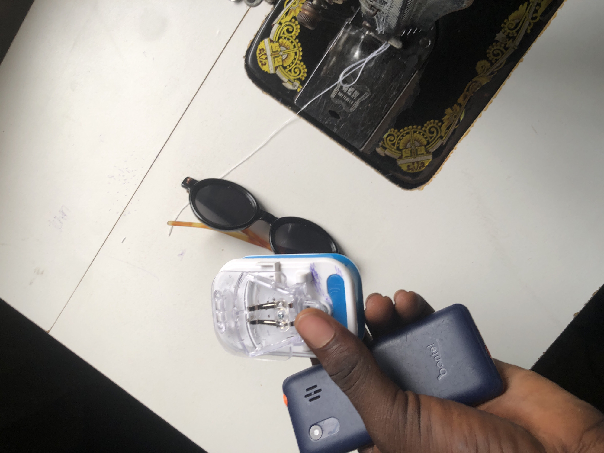 A hand is holding a dark blue Bontel mobile phone and a universal mobile phone charger. Next to the charger are a pair of black-rimmed sunglasses with orange arms. In the background, a vintage black sewing machine with gold decorative elements sits on a white surface, possibly a table. A white thread is visible on the sewing machine and extending across the surface. The scene is likely in Ado, Nigeria, given the presence of a Bontel phone, a brand popular in that region.