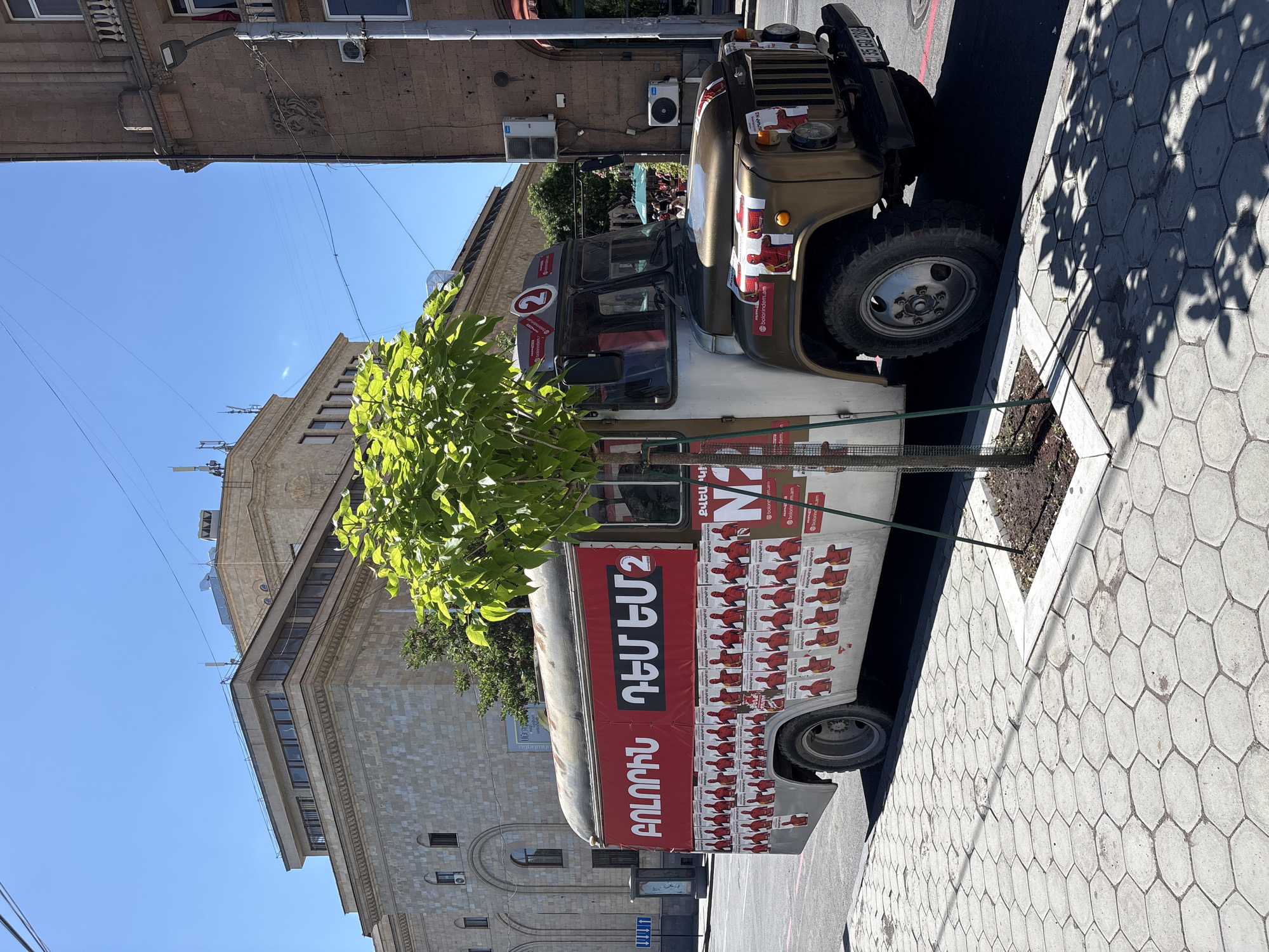 The image depicts a vehicle parked on a city street, likely in Yerevan, Armenia, based on the language on the vehicle's advertising. The vehicle is a light-colored truck or van, heavily stickered with political advertisements featuring a red background. The lettering and images on the stickers are in Armenian and likely promote a political campaign. The building in the background appears to be a governmental or civic structure, constructed of light-colored stone or concrete. A small, recently planted tree is situated between the vehicle and the sidewalk. The sidewalk is paved with hexagonal pavers, and shadows from another tree are cast upon it. The weather appears to be sunny, with a clear blue sky visible above the buildings.